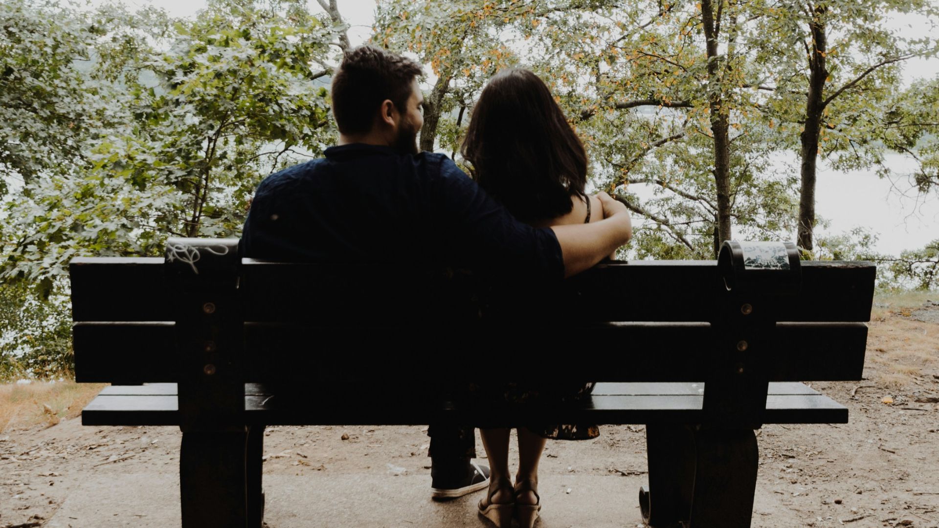 man looking to woman sitting on black wooden bench in front of tall trees during daytime