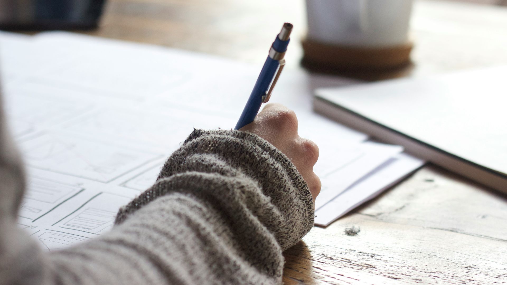 person writing on brown wooden table near white ceramic mug