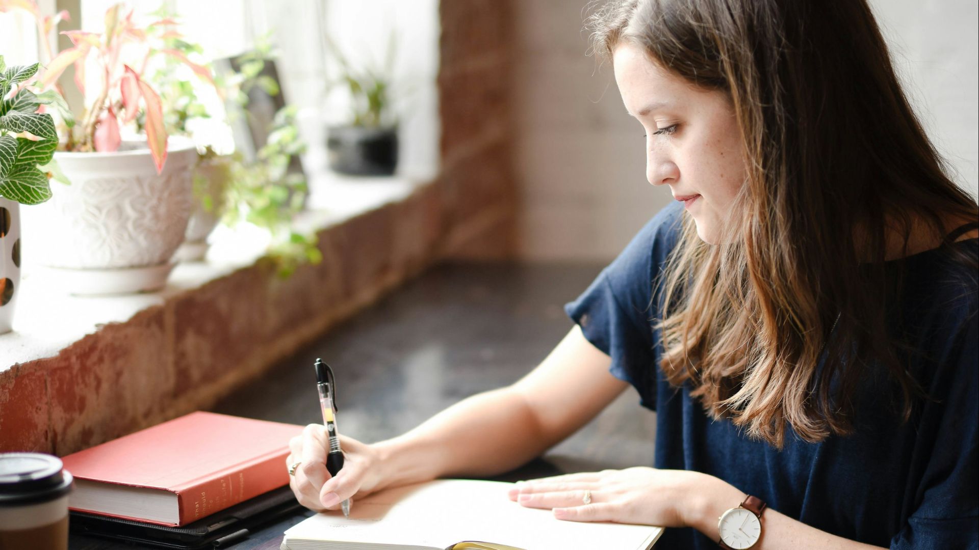 woman sitting in front of black table writing on white book near window