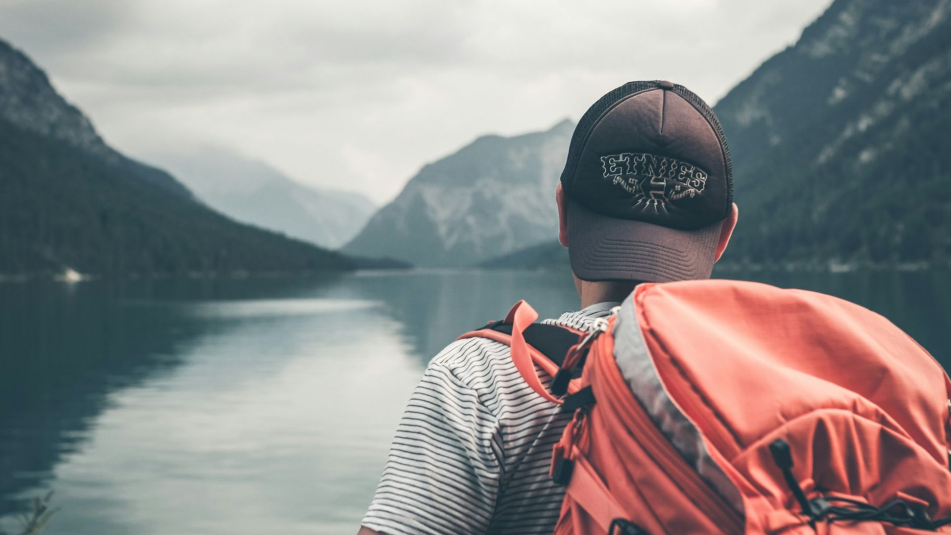 man with red hiking backpack facing body of water and mountains at daytime