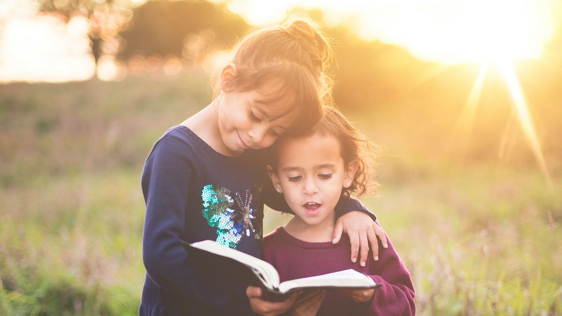 girl's left hand wrap around toddler while reading book during golden hour