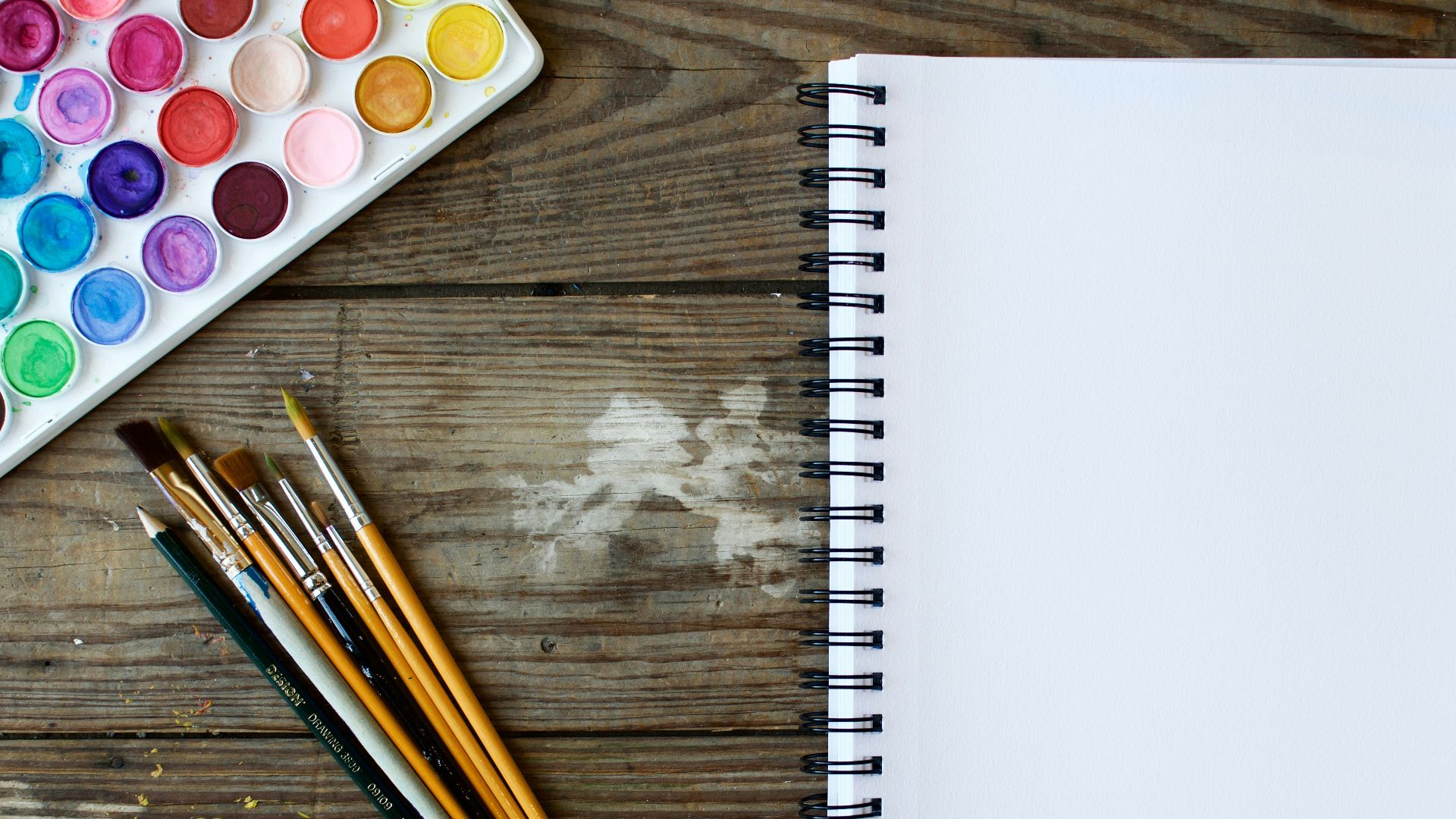 paint brushes next to drawing book and water color palette