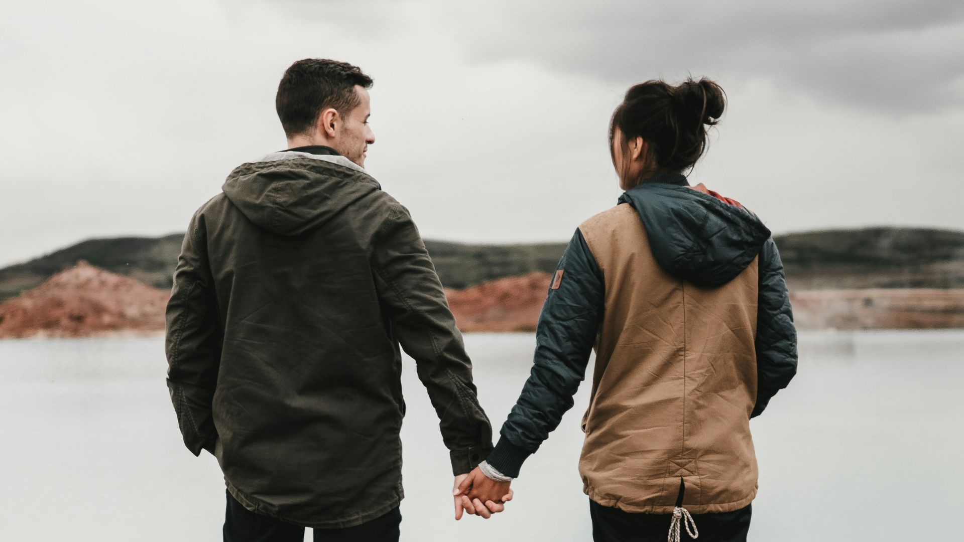 couple holding hand front of body of calm water with mountain distance