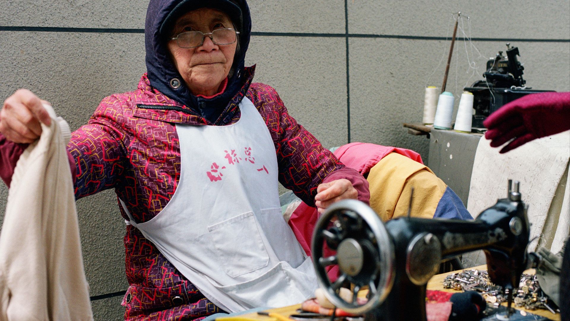 An elderly woman sews at her outdoor workstation.