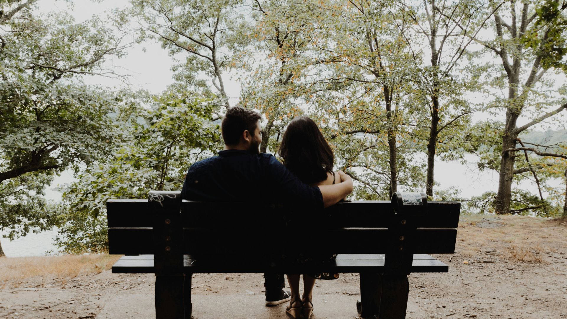 man looking to woman sitting on black wooden bench in front of tall trees during daytime