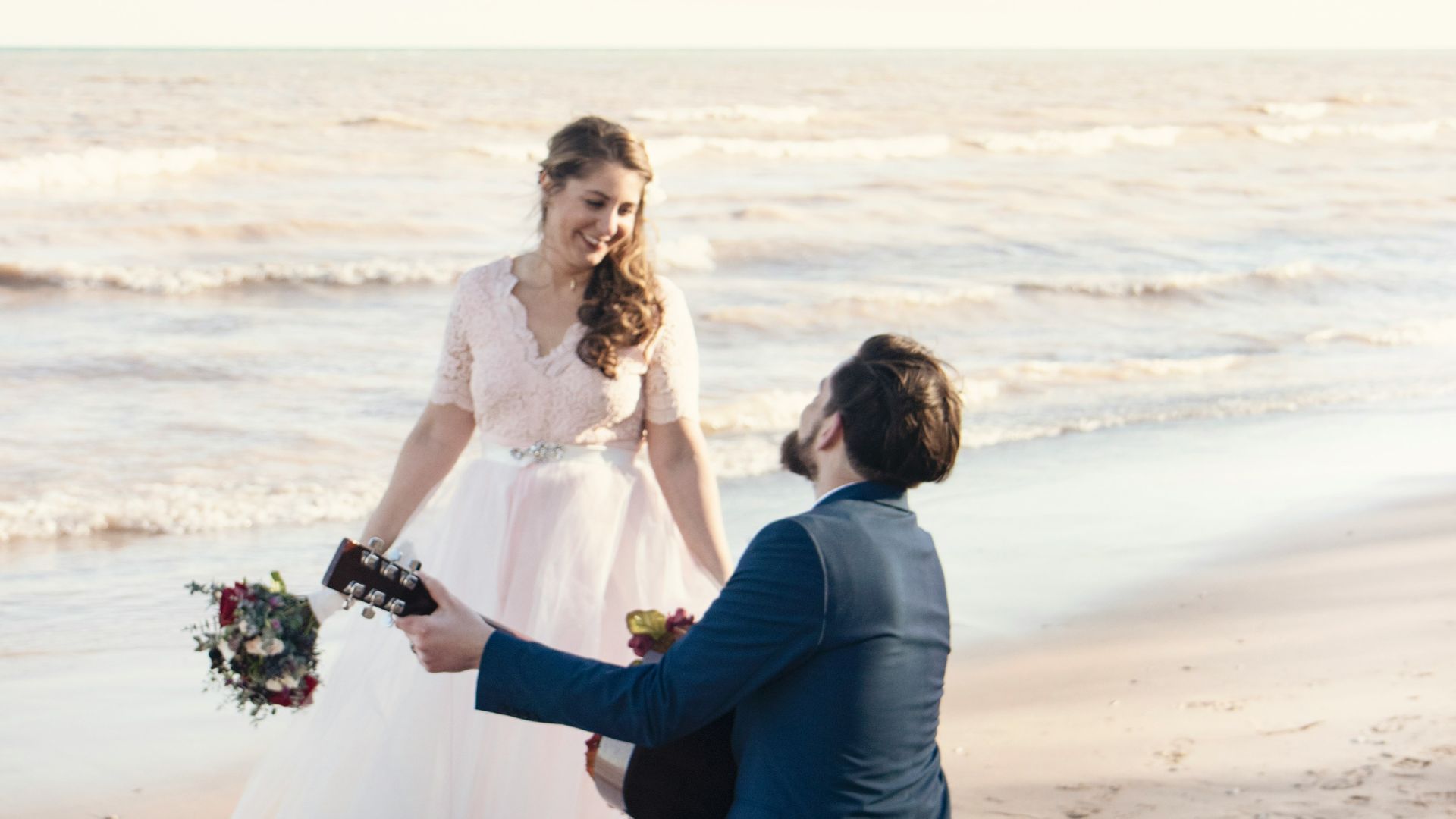 man kneeling on seashore and playing guitar in front of woman at daytime