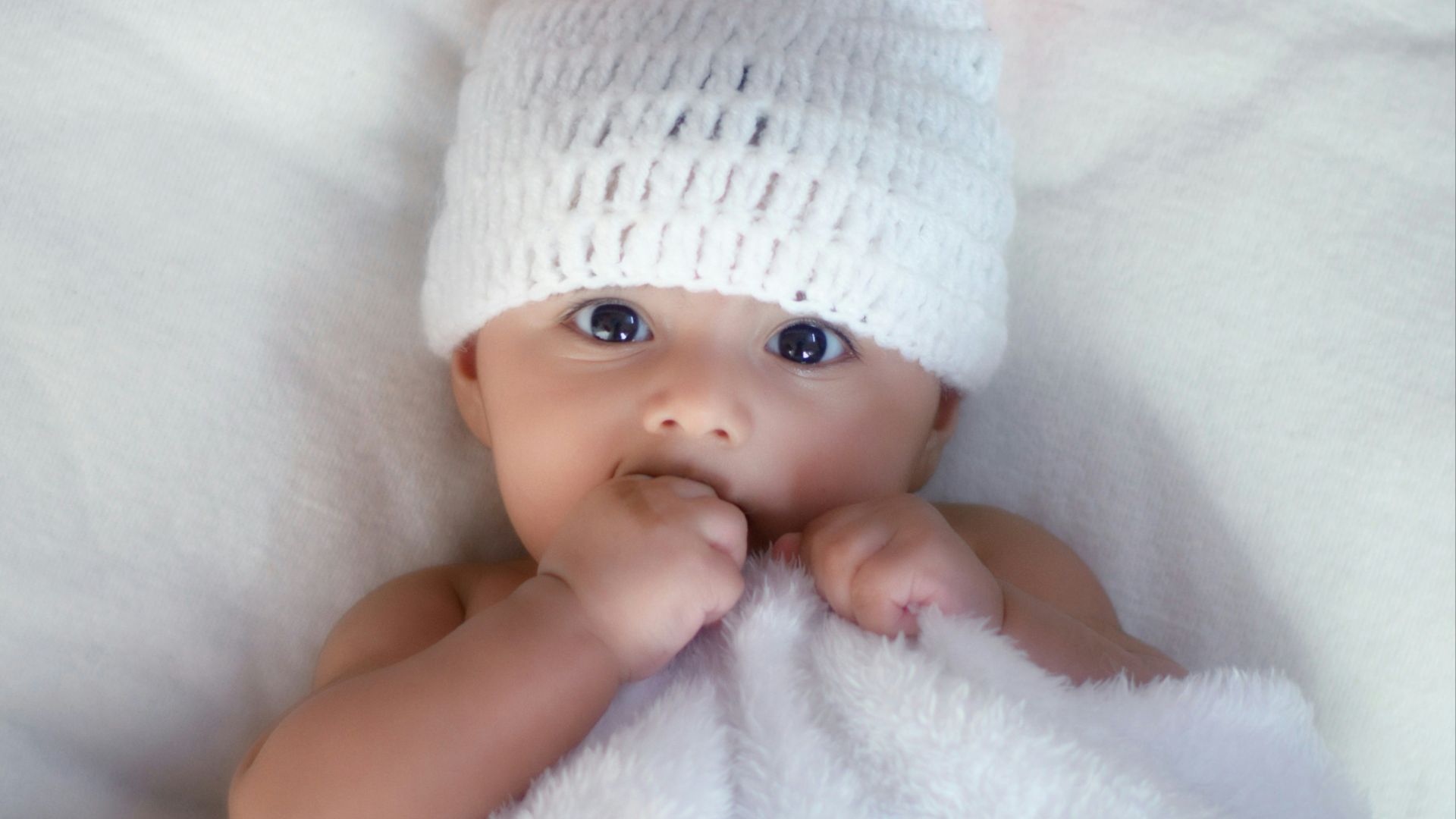 baby in white knit cap lying on white textile