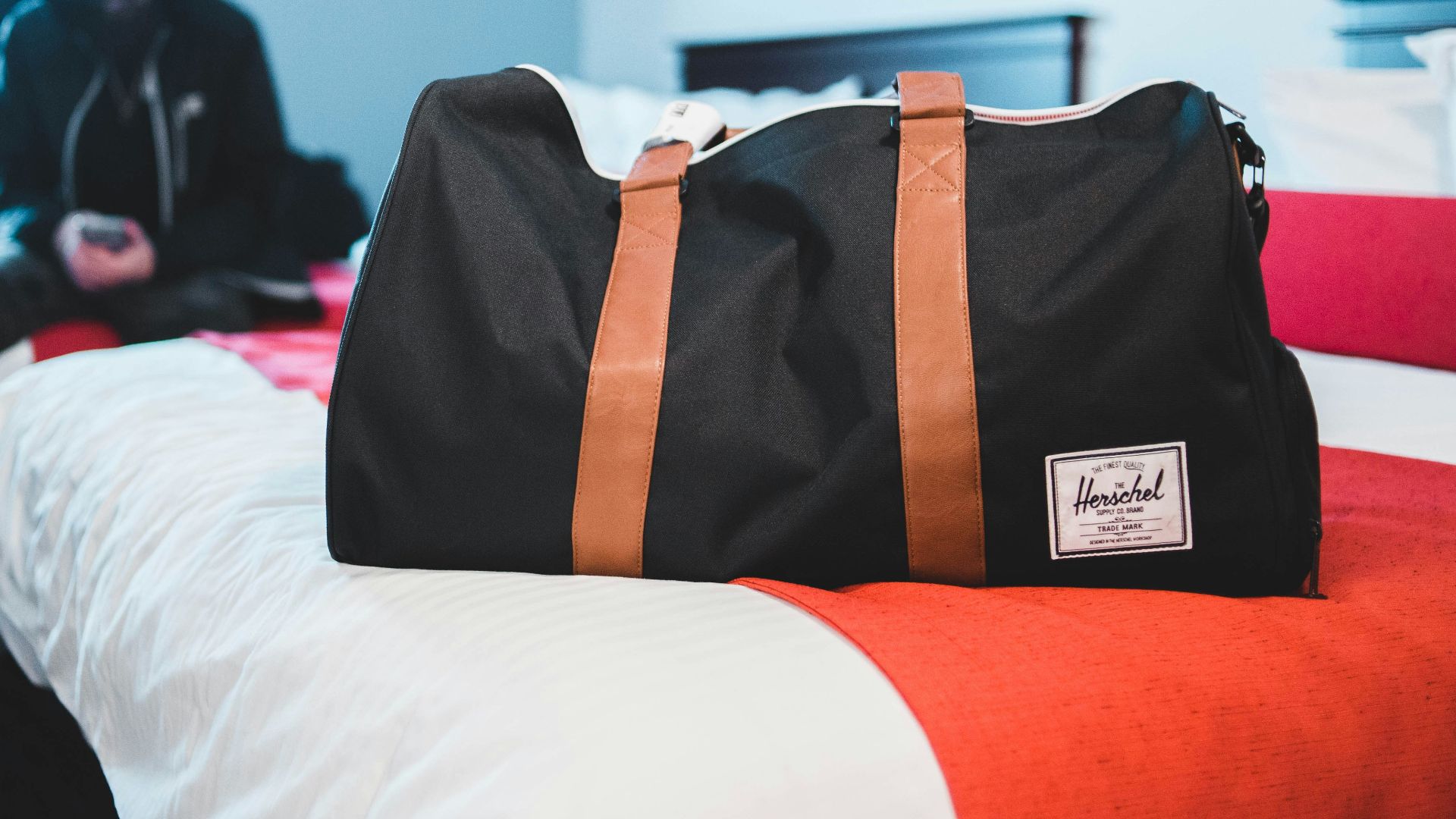 man in black hooded jacket sitting on bed near black and brown leather tote bag