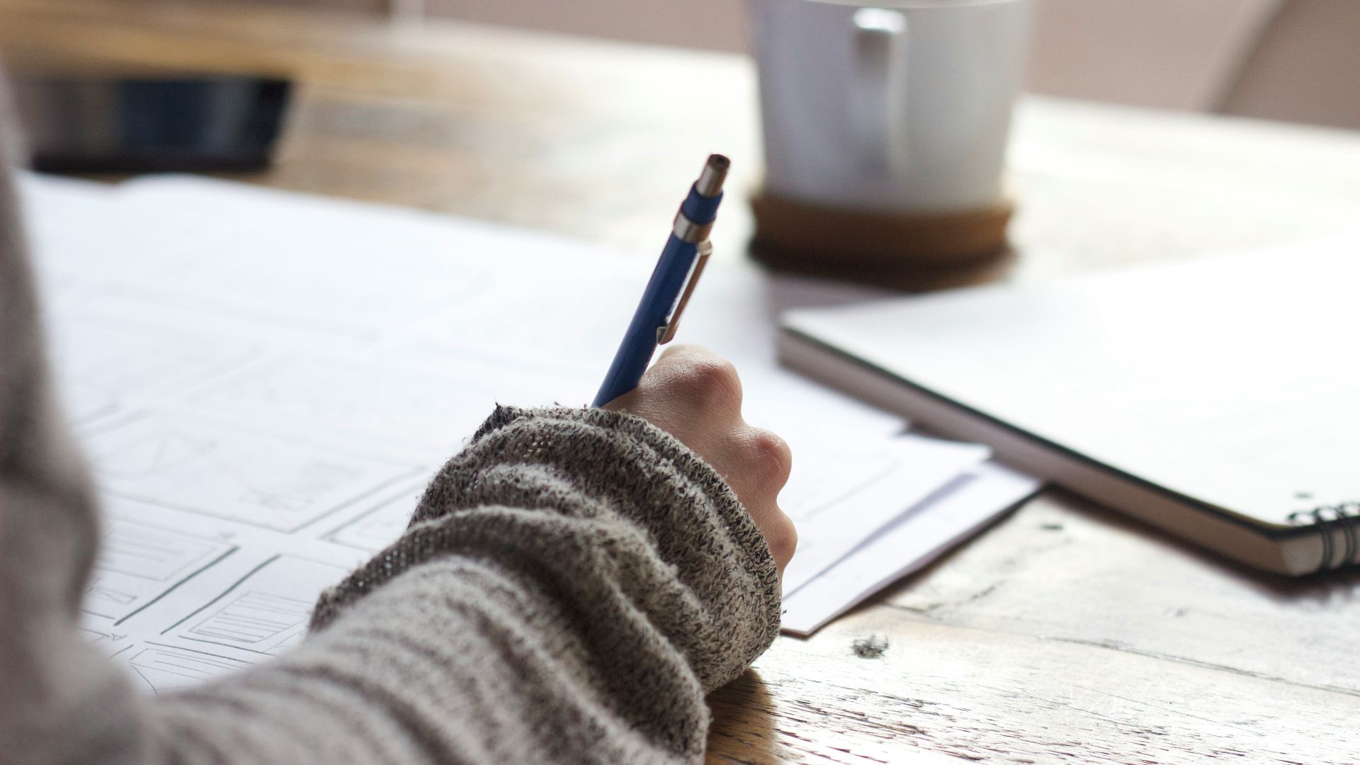 person writing on brown wooden table near white ceramic mug