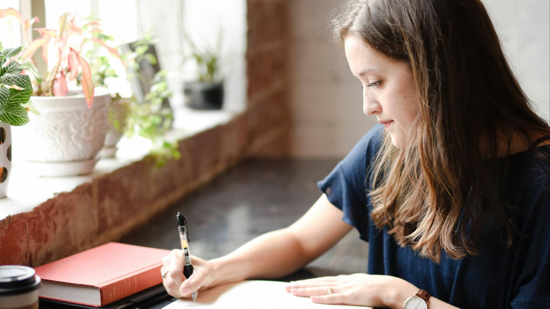 woman sitting in front of black table writing on white book near window