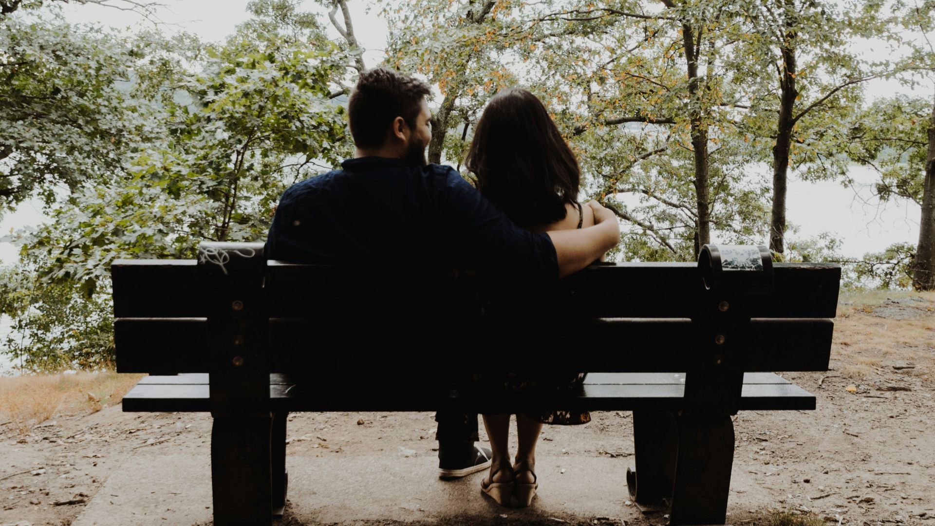 man looking to woman sitting on black wooden bench in front of tall trees during daytime