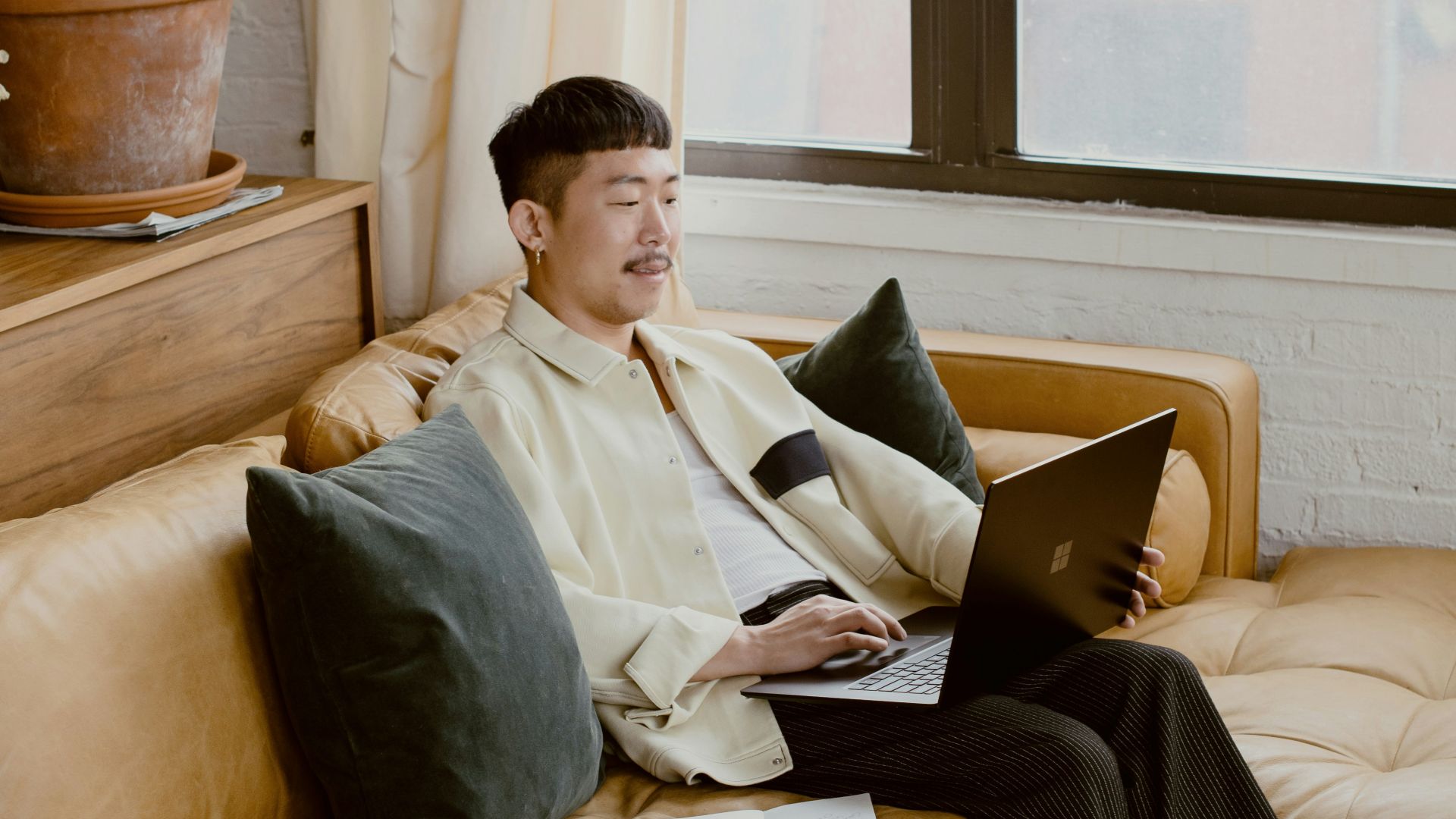 man in white dress shirt sitting on white couch using black Surface laptop