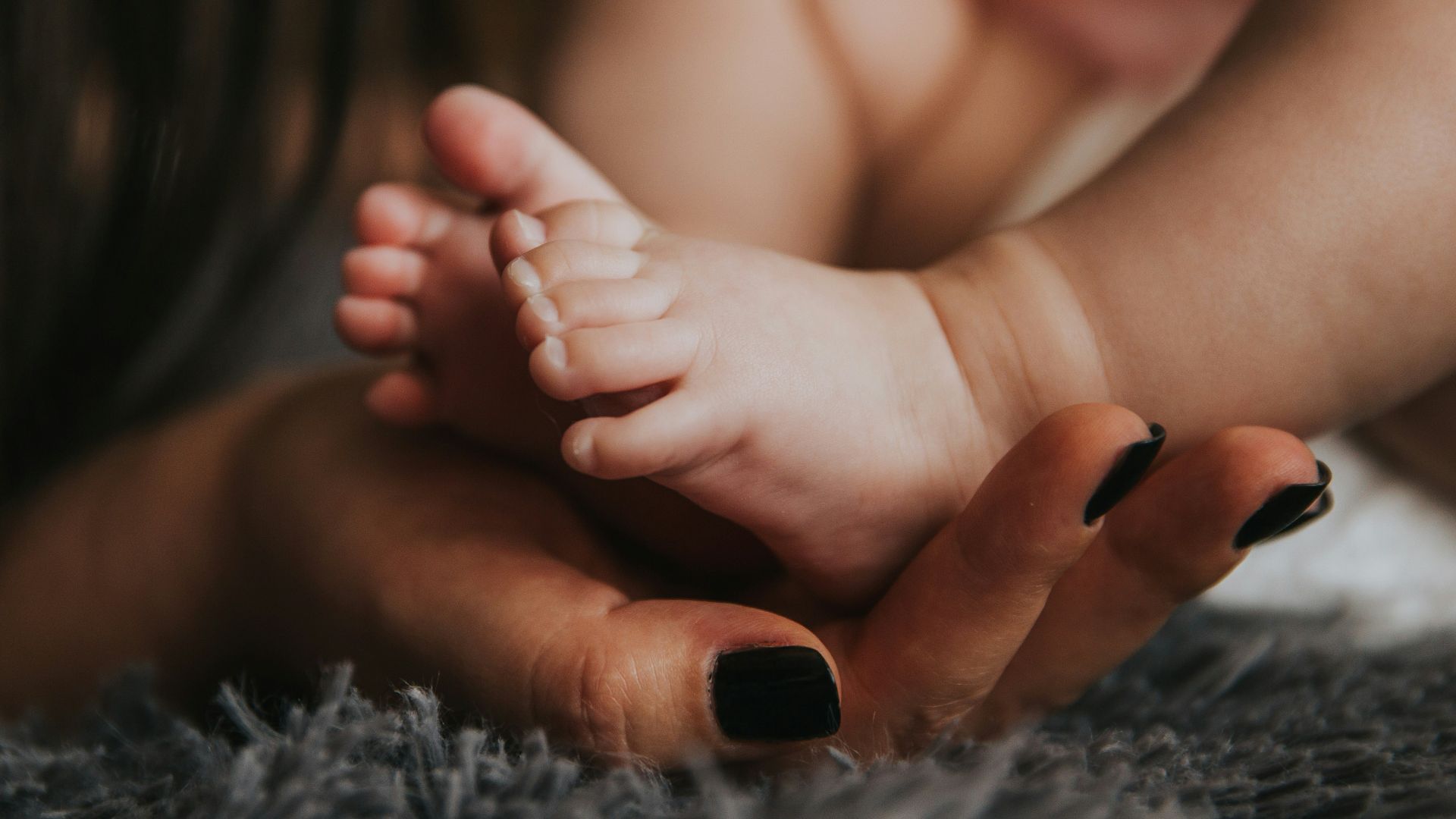 Infant's feet being held by a woman's hand with painted and manicured hands resting on a gray blanket