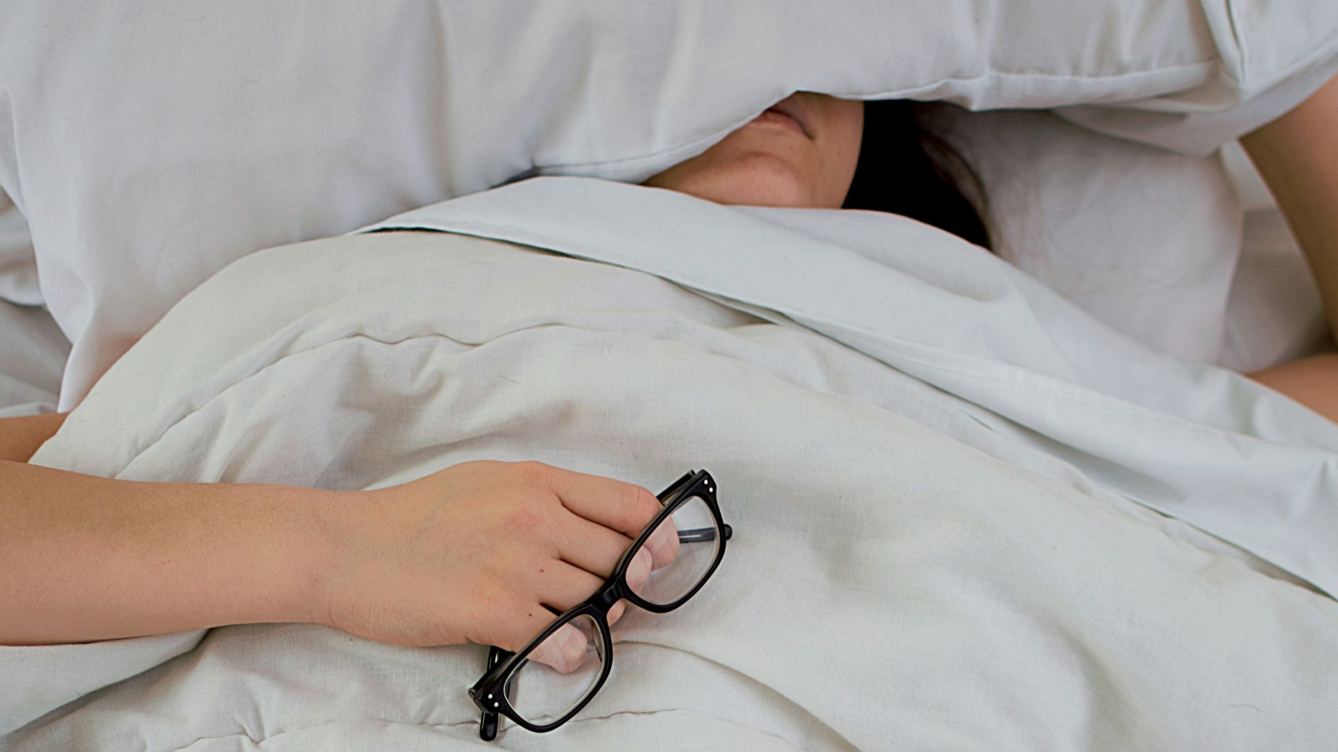 person lying on bed while covering face with pillow and holding eyeglasses