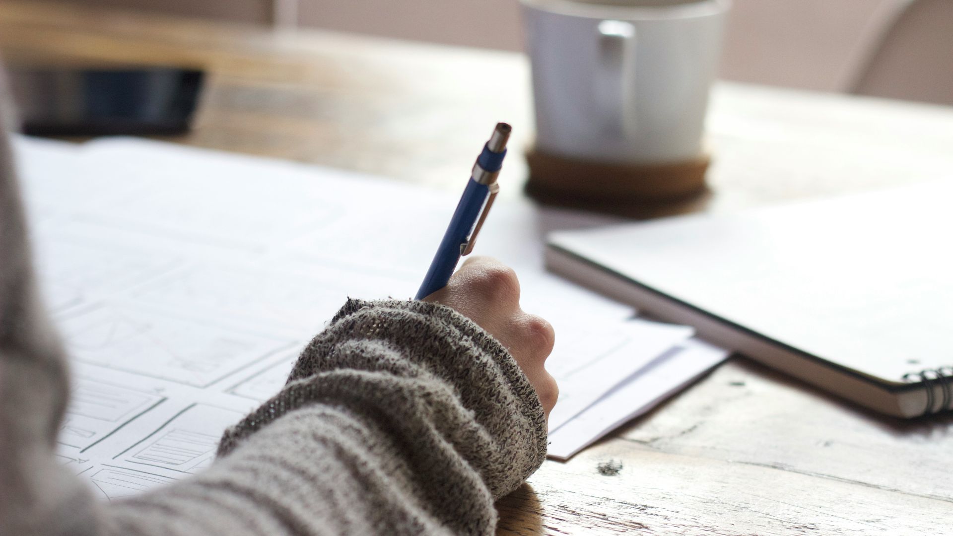 person writing on brown wooden table near white ceramic mug