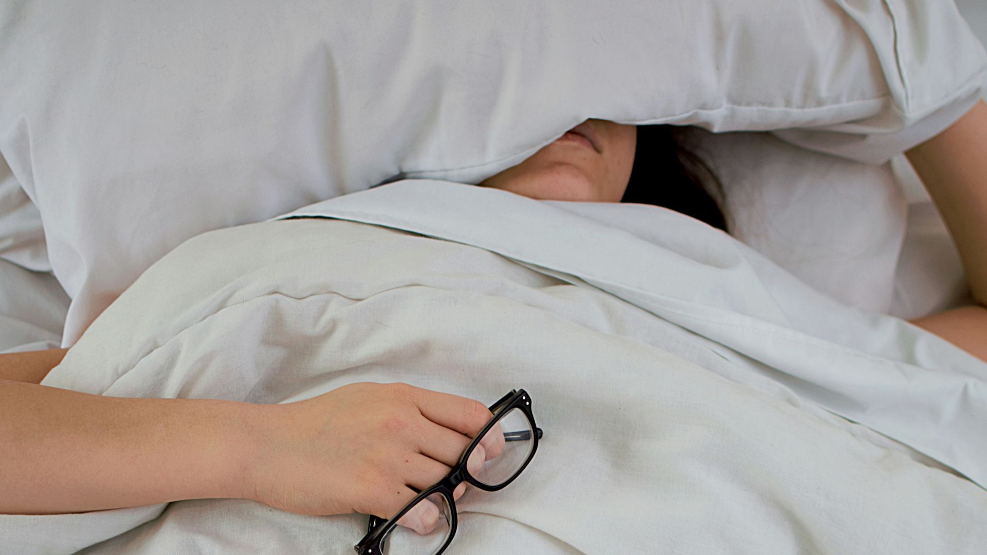 person lying on bed while covering face with pillow and holding eyeglasses