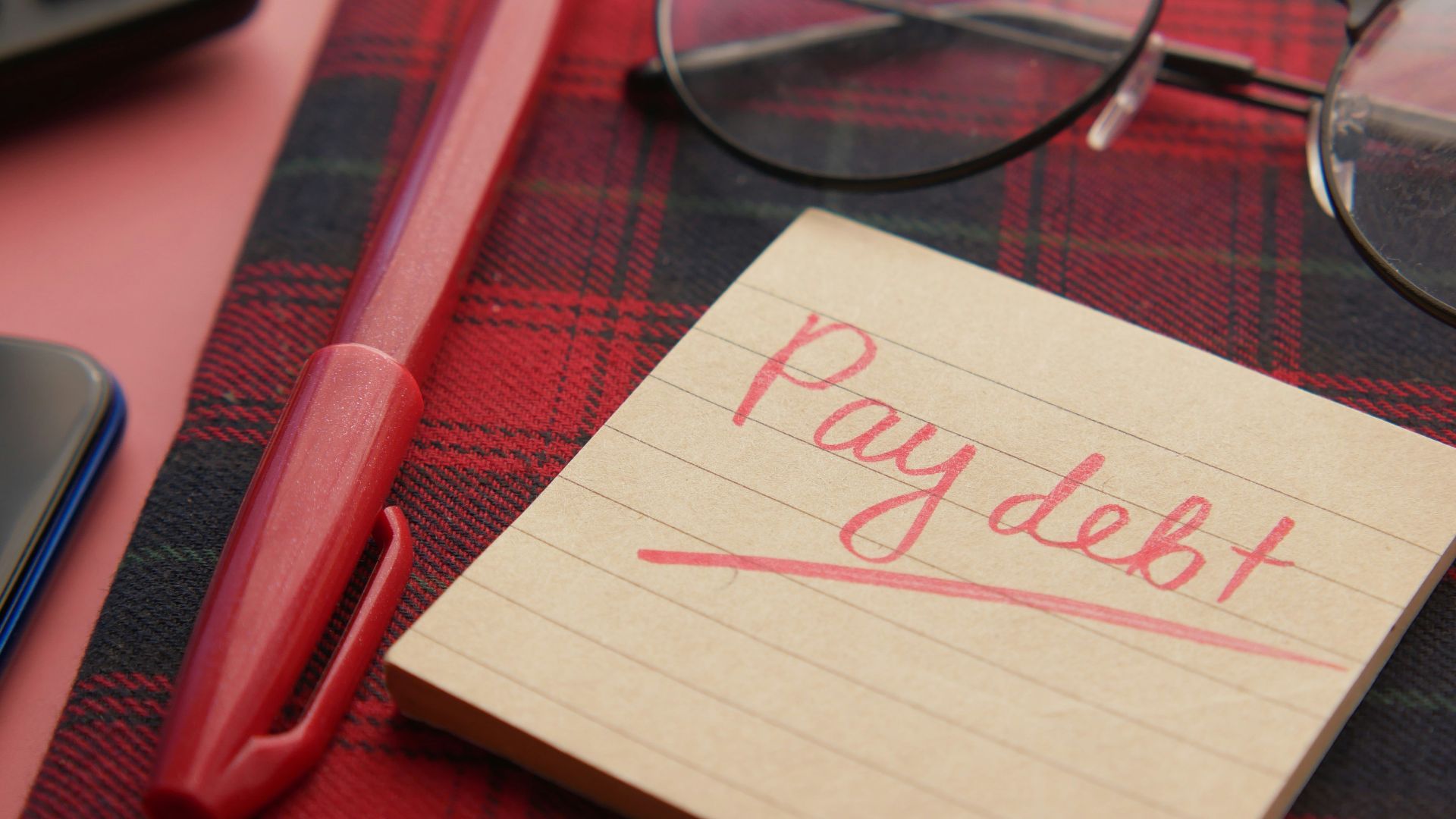 a note that says pay debt next to a pen and glasses