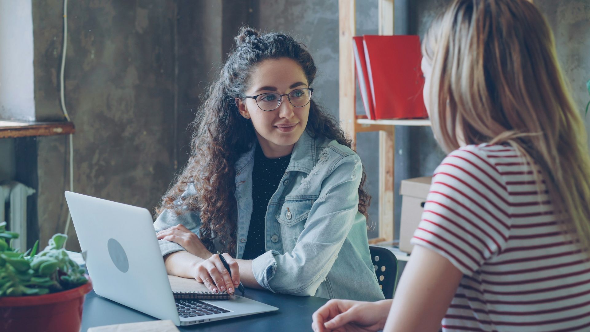Two women are having a conversation at a desk.