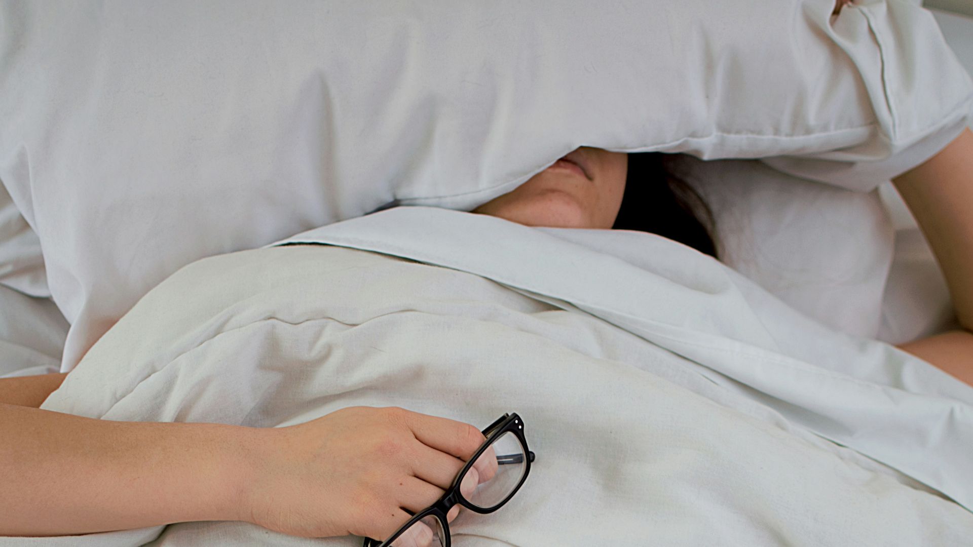 person lying on bed while covering face with pillow and holding eyeglasses