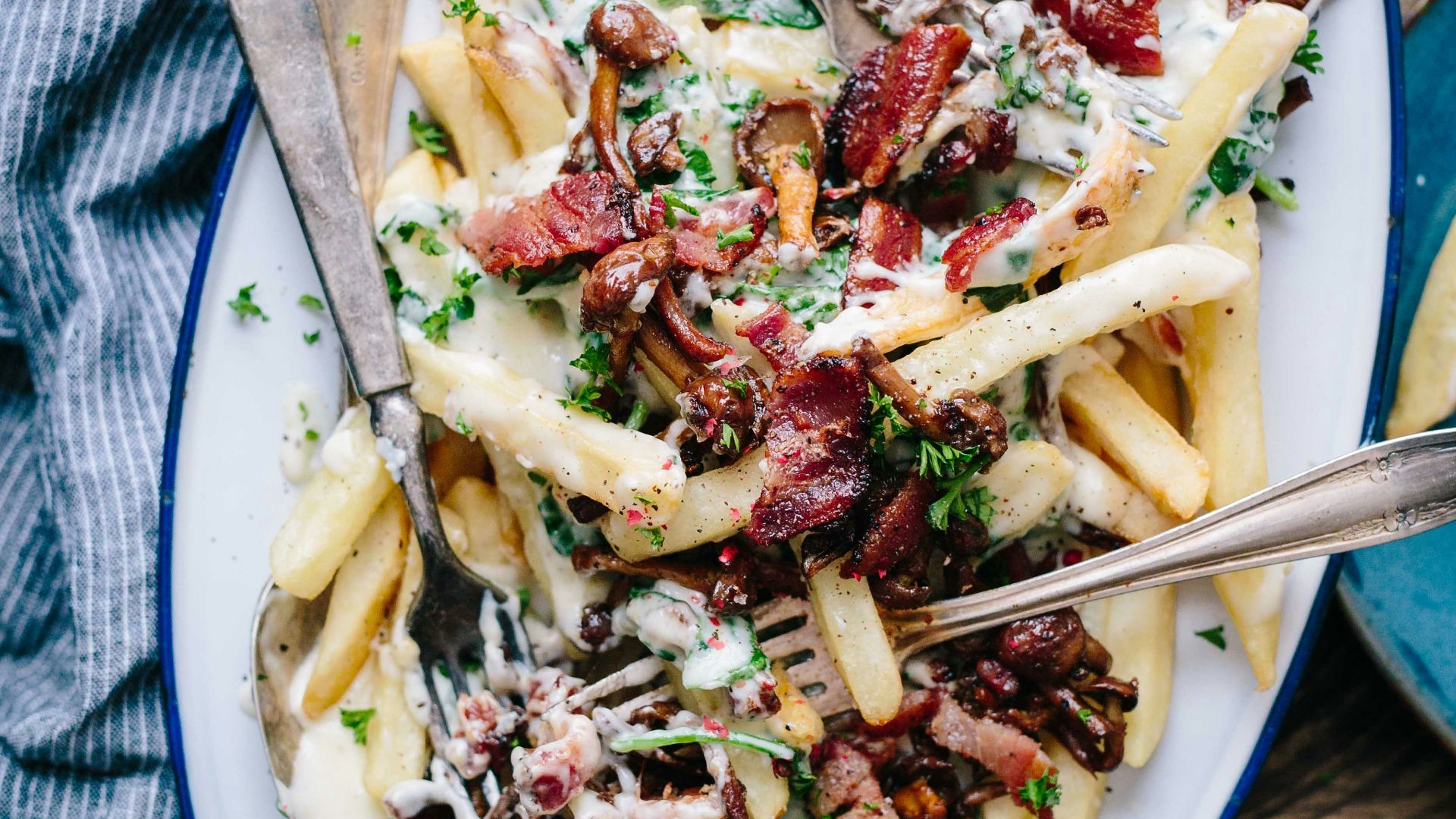 bacon strips and melted cheese topped fries on oval white and blue platter with gray stainless steel forks