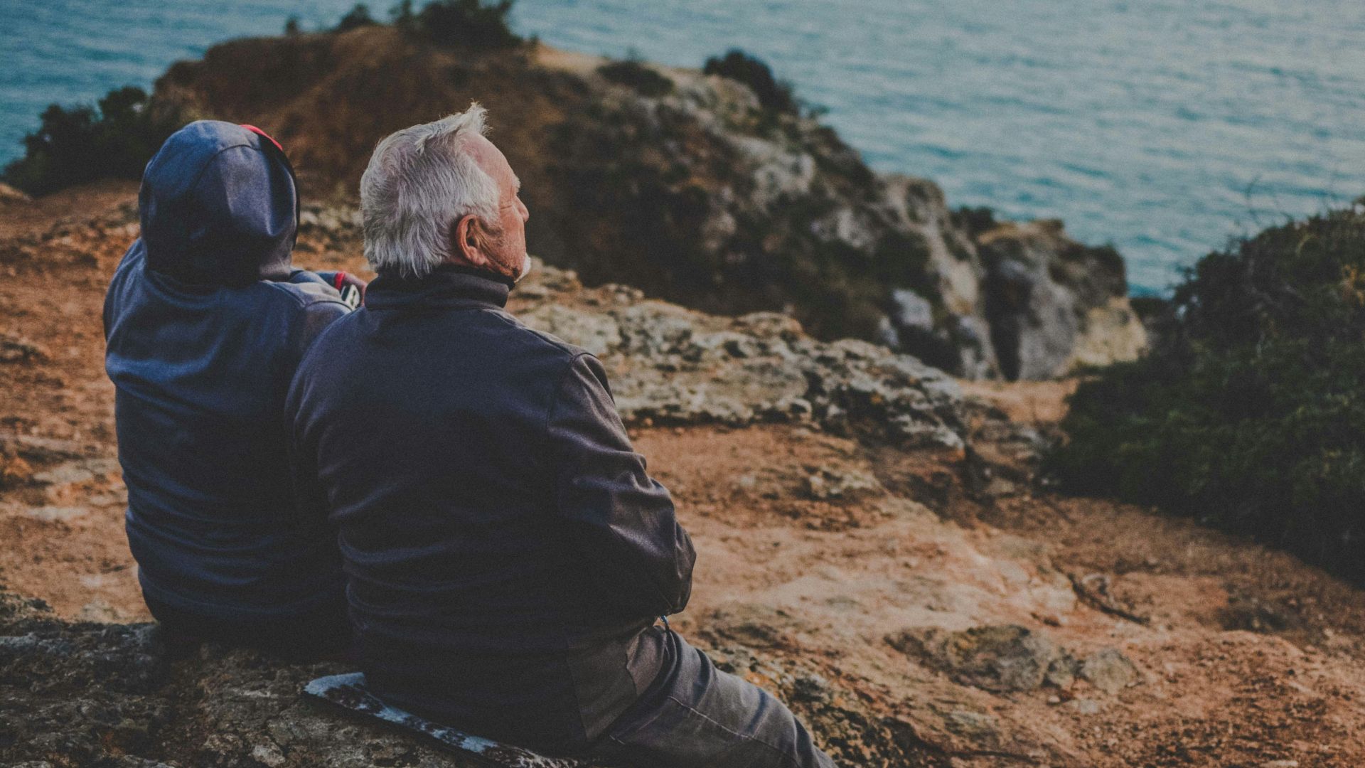 two person sitting on rock staring at body of water during daytime