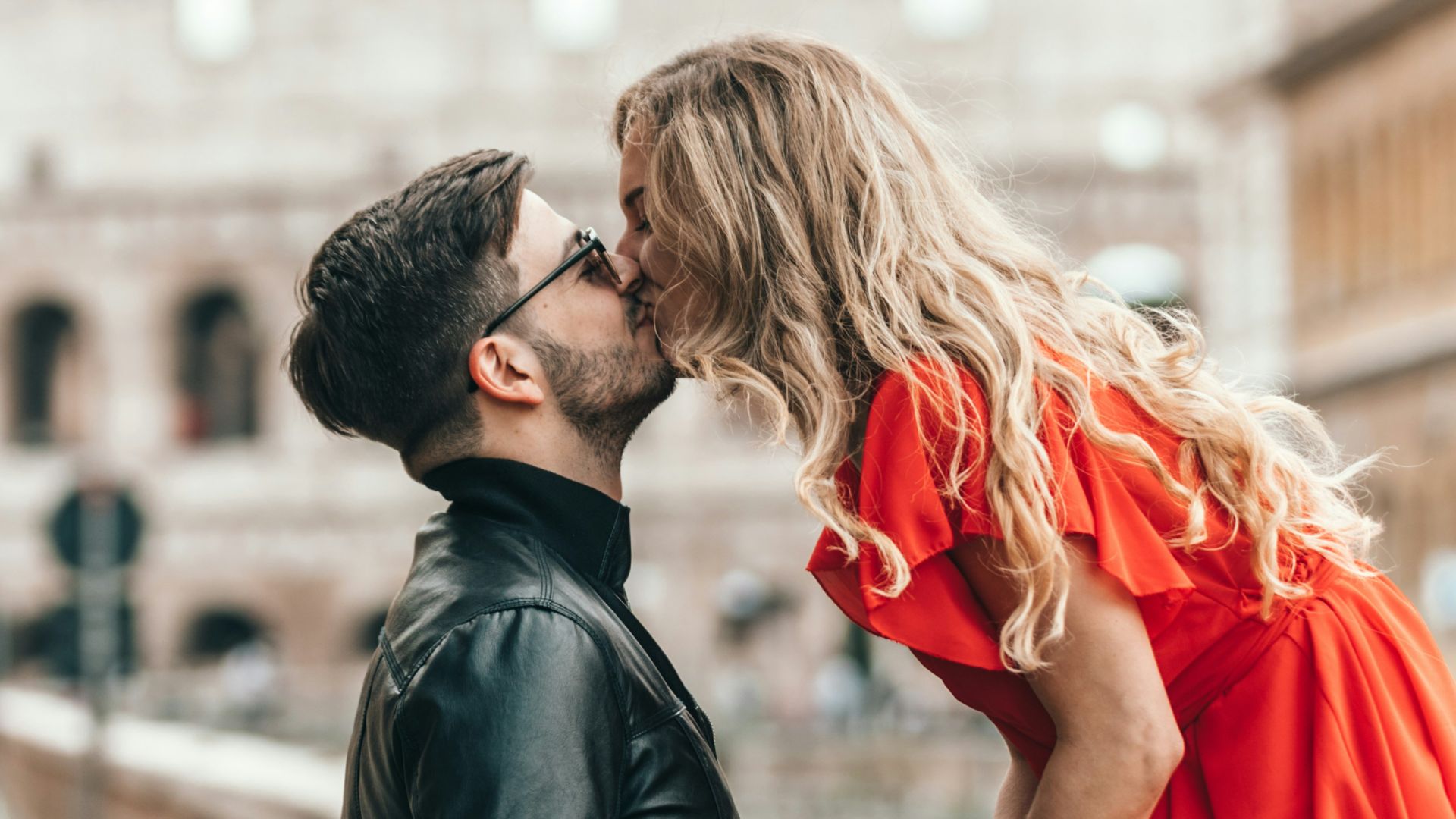 man standing and kissing woman sitting on concrete wall