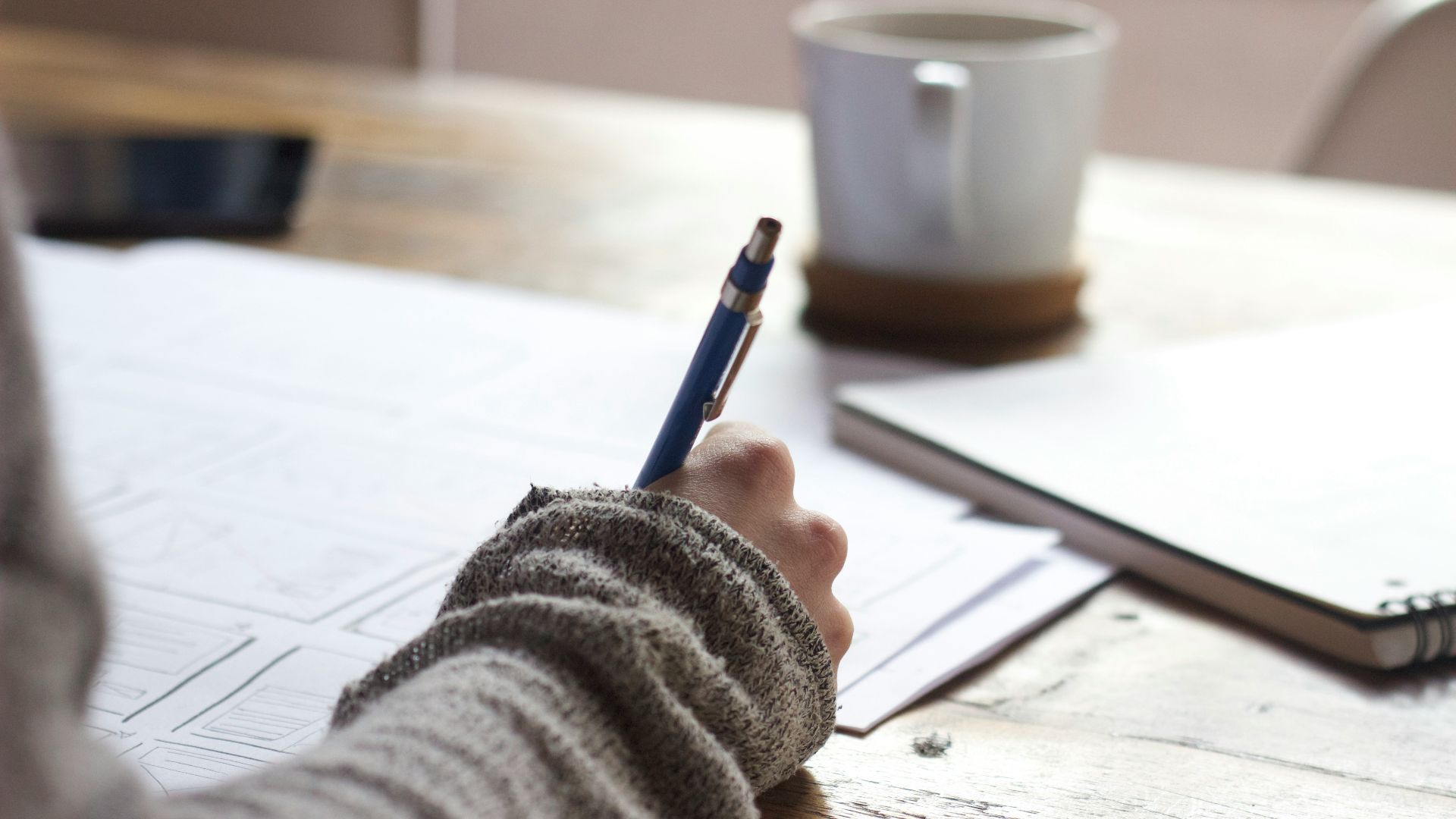person writing on brown wooden table near white ceramic mug
