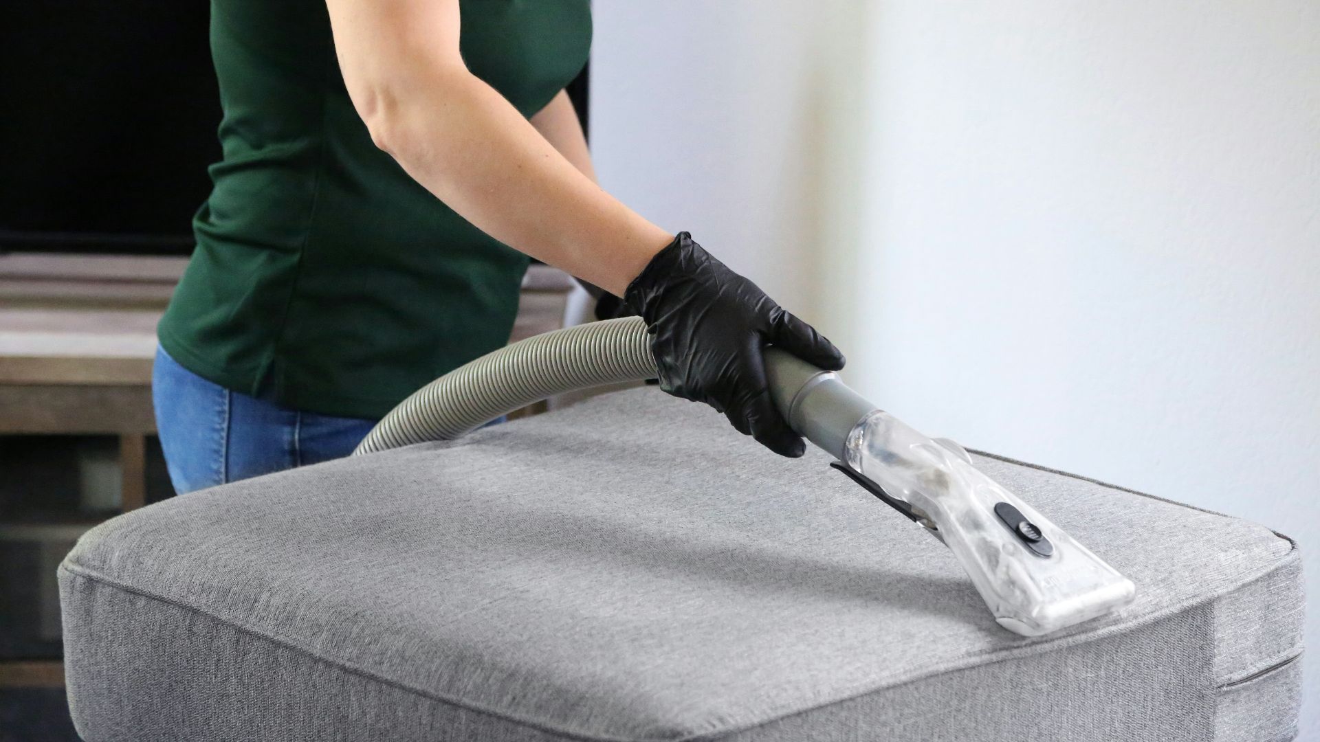a woman in a green shirt and black gloves vacuuming a gray ottoman