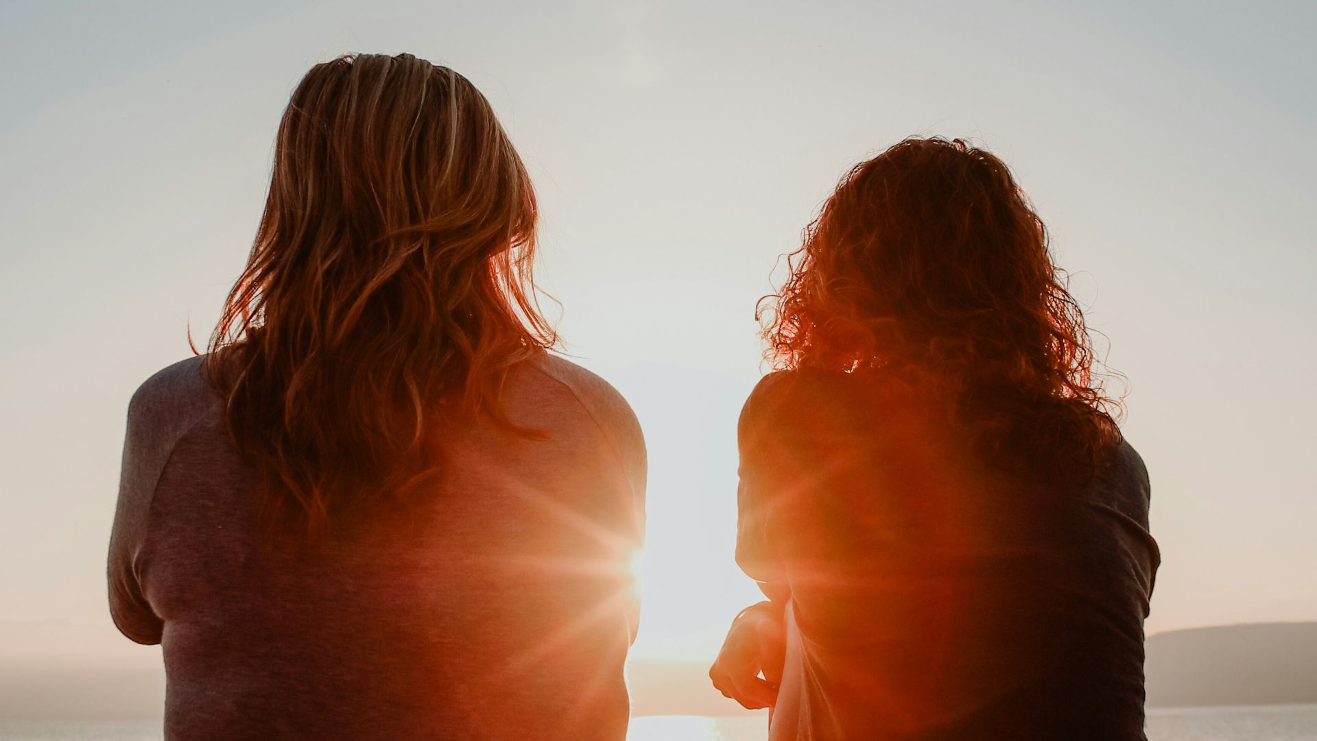 two woman sitting on beach sand while facing sunlight
