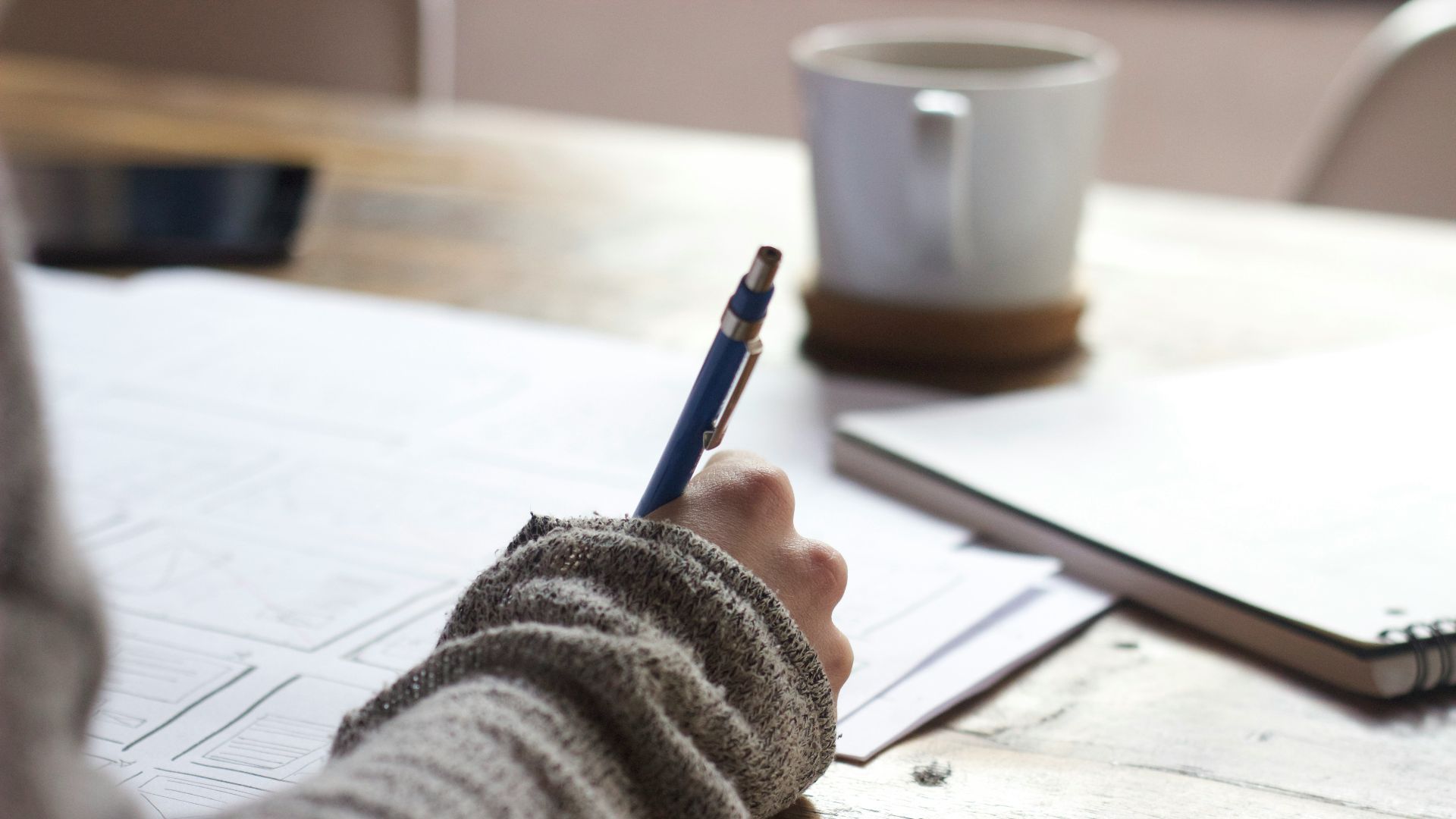 person writing on brown wooden table near white ceramic mug