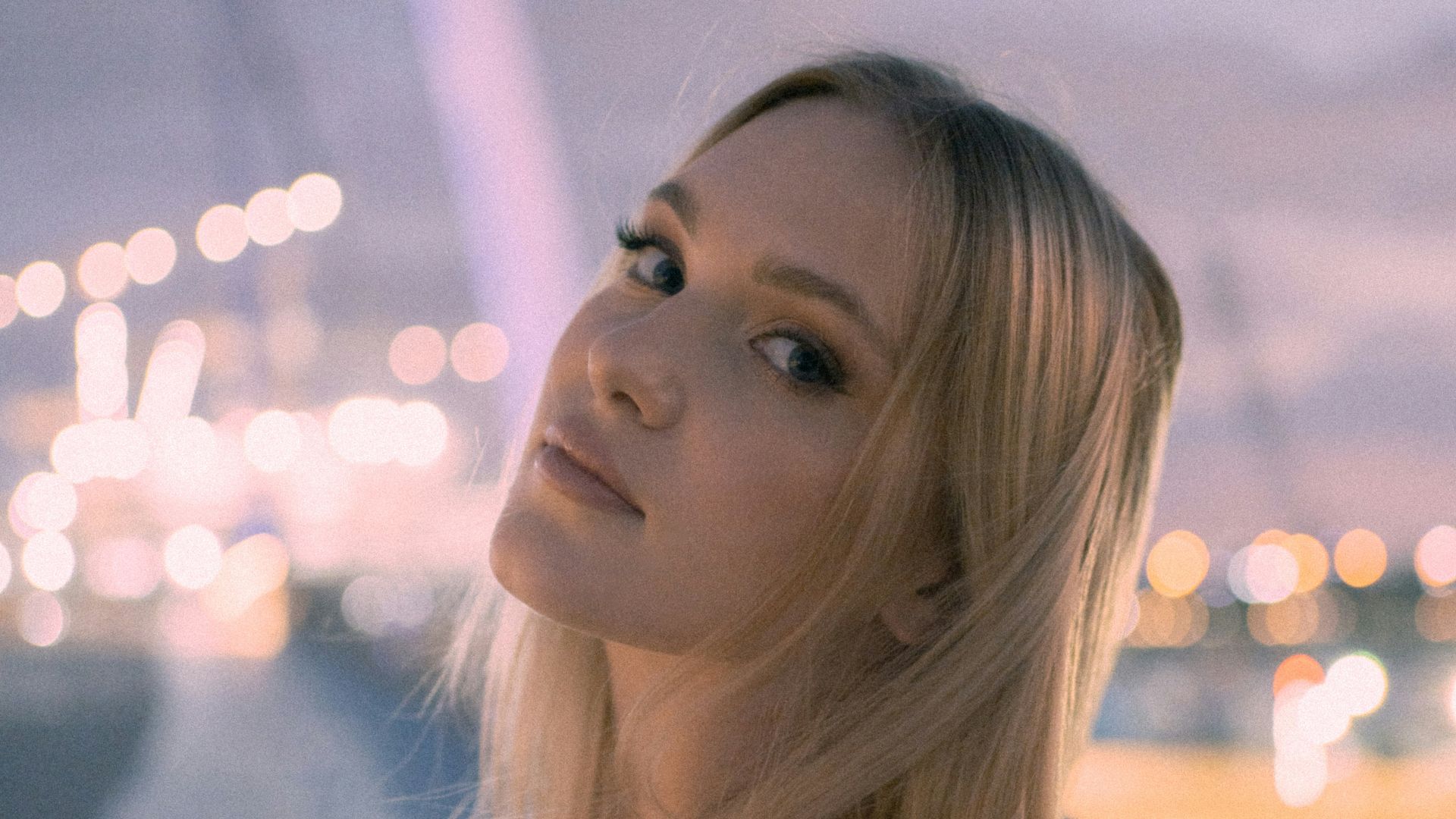 Blonde woman poses by a bridge at dusk.