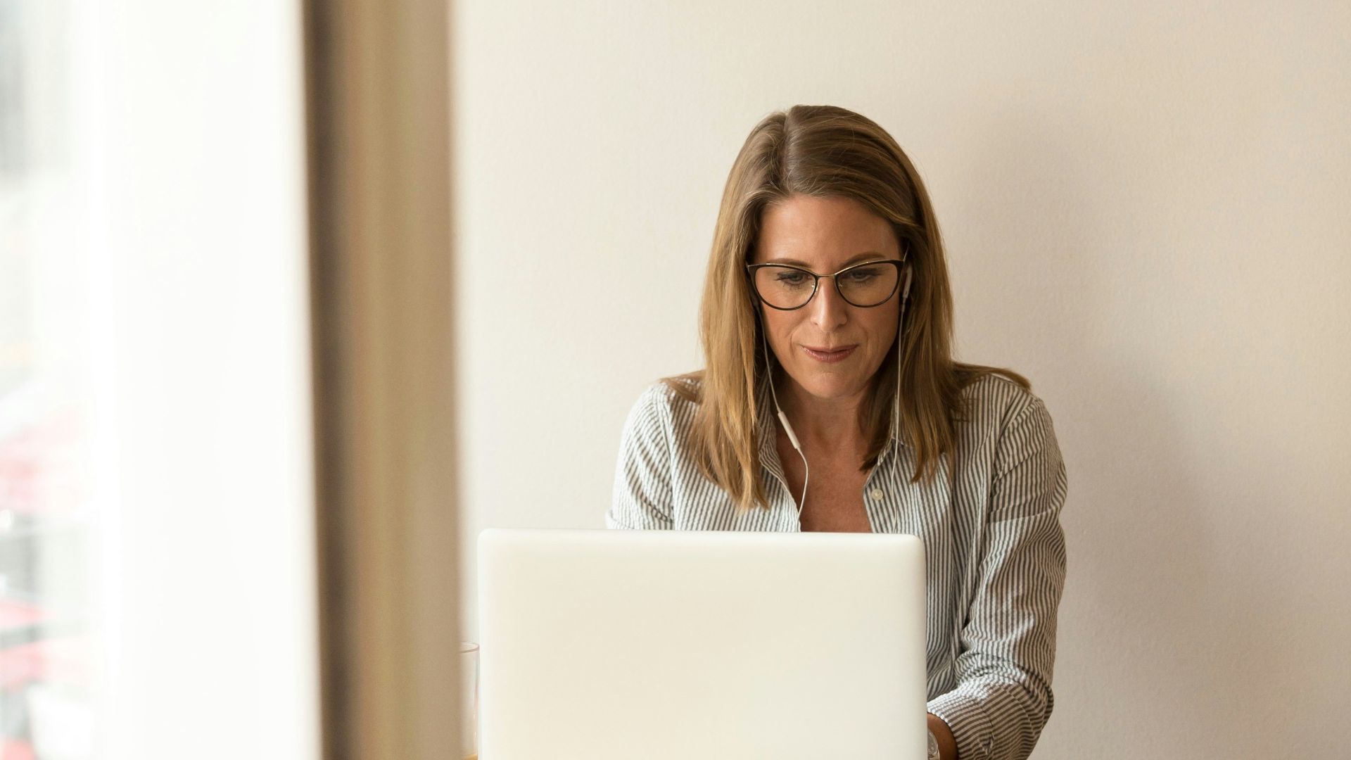 woman wearing grey striped dress shirt sitting down near brown wooden table in front of white laptop computer