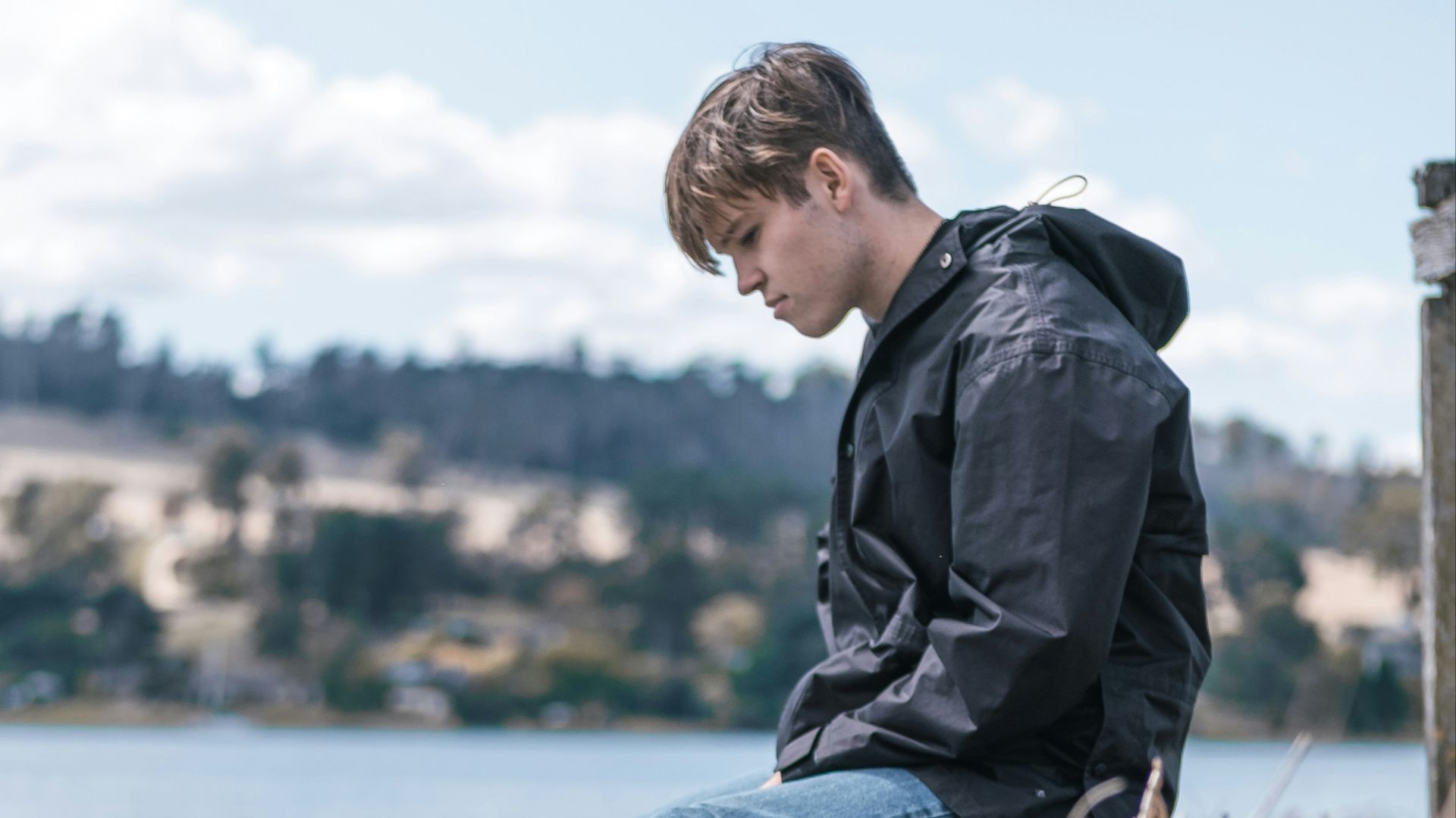 man in black jacket and blue denim jeans sitting on brown wooden log during daytime