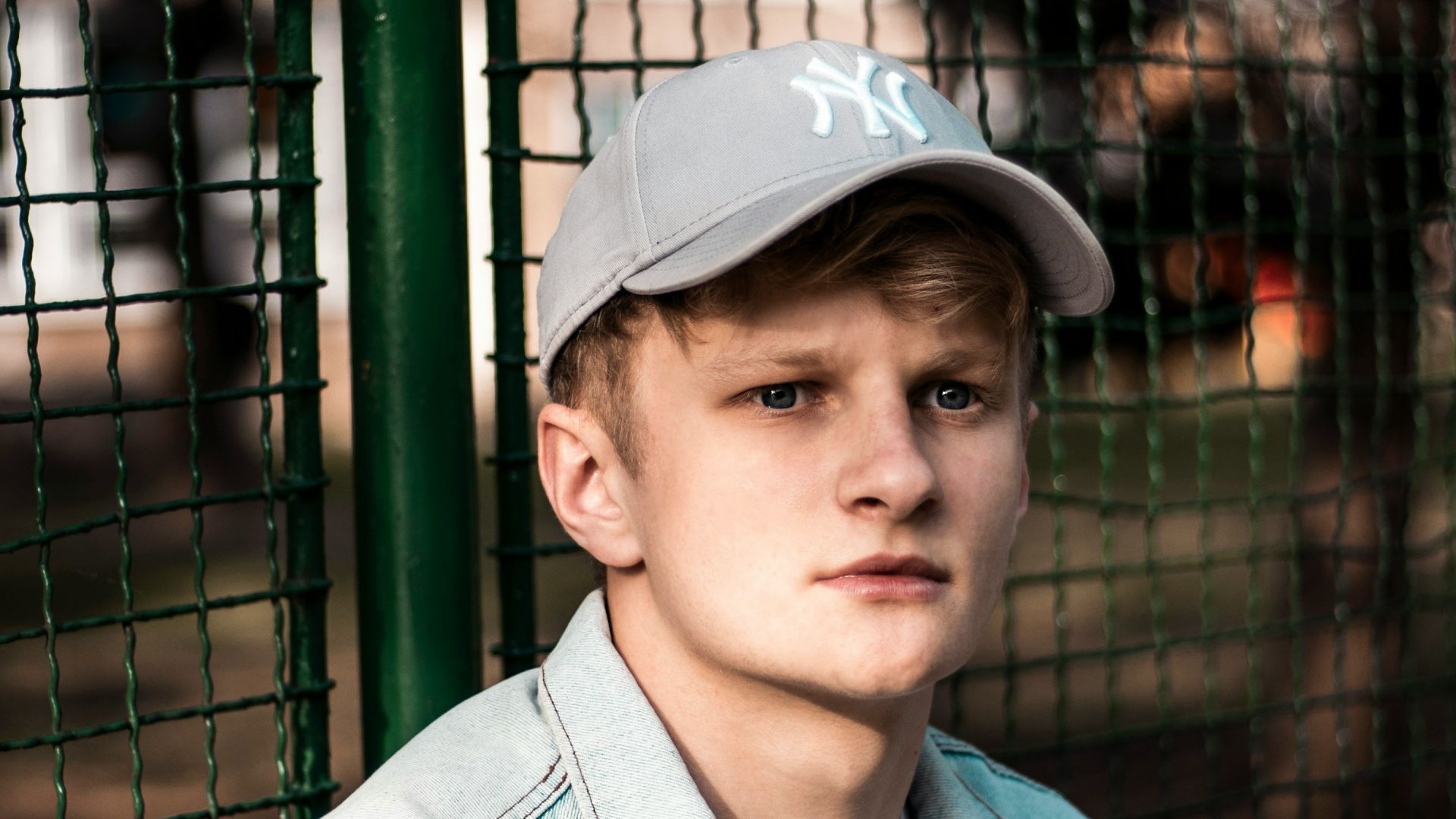 man wearing blue button-up denim jacket sitting beside green fence