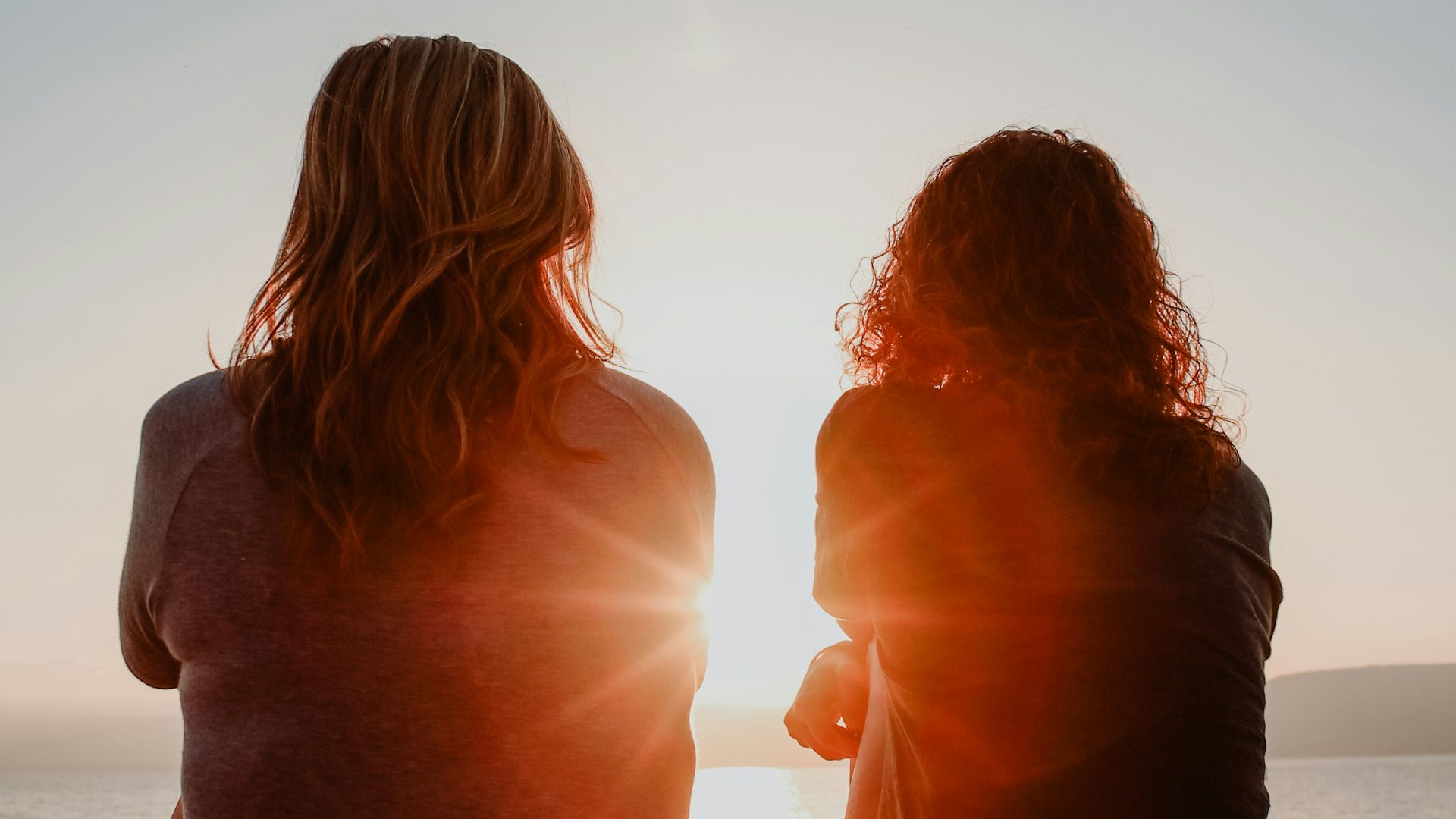 two woman sitting on beach sand while facing sunlight