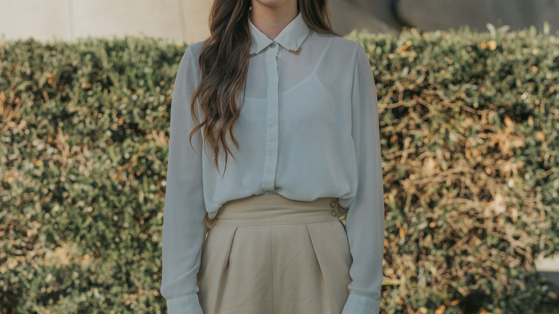 woman wearing gray button-up long-sleeved shirt and brown pants standing in front of bushes