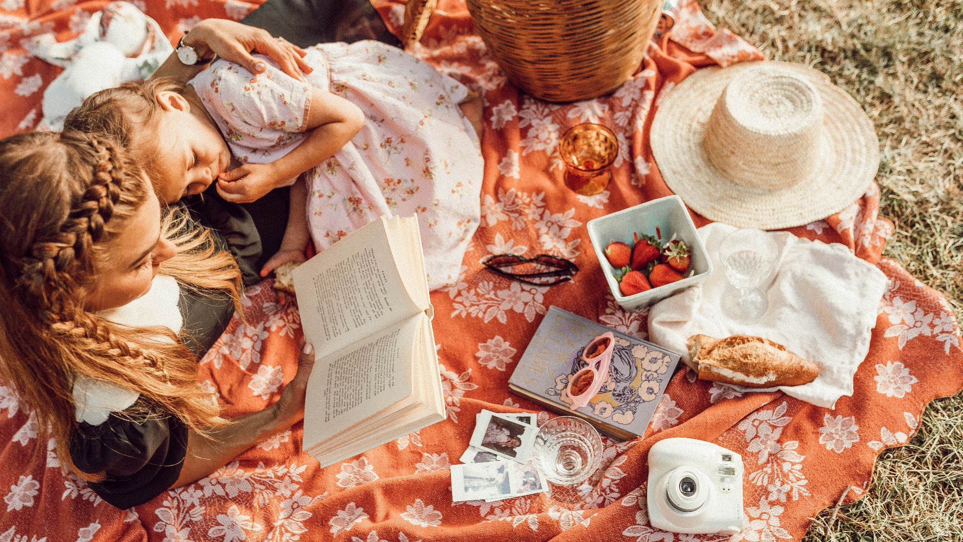 woman reading with girl while lying on orange and white floral picnic mat