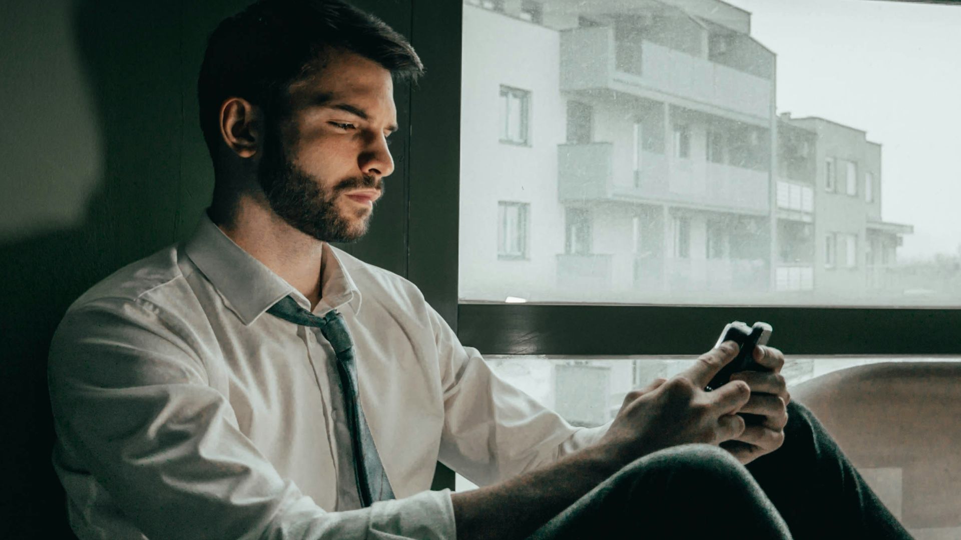 a man sitting on a window sill looking at his cell phone