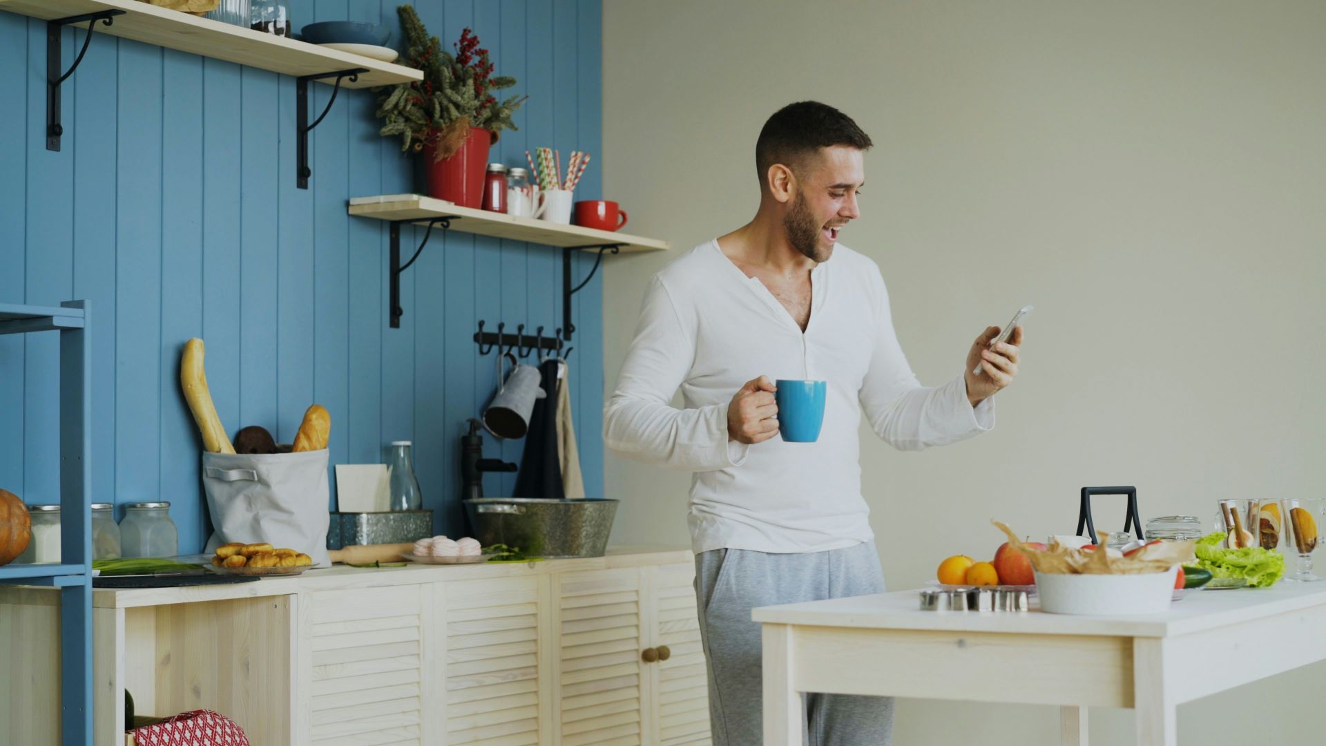 Man holding coffee cup and looking at phone in kitchen.