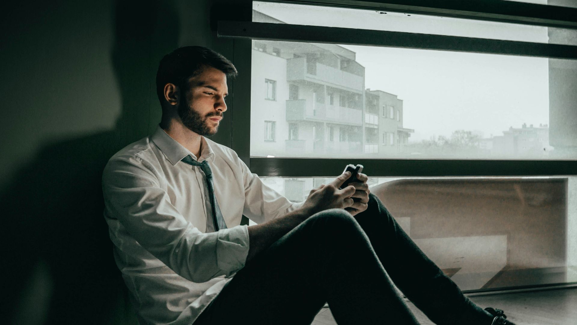 a man sitting on a window sill looking at his cell phone