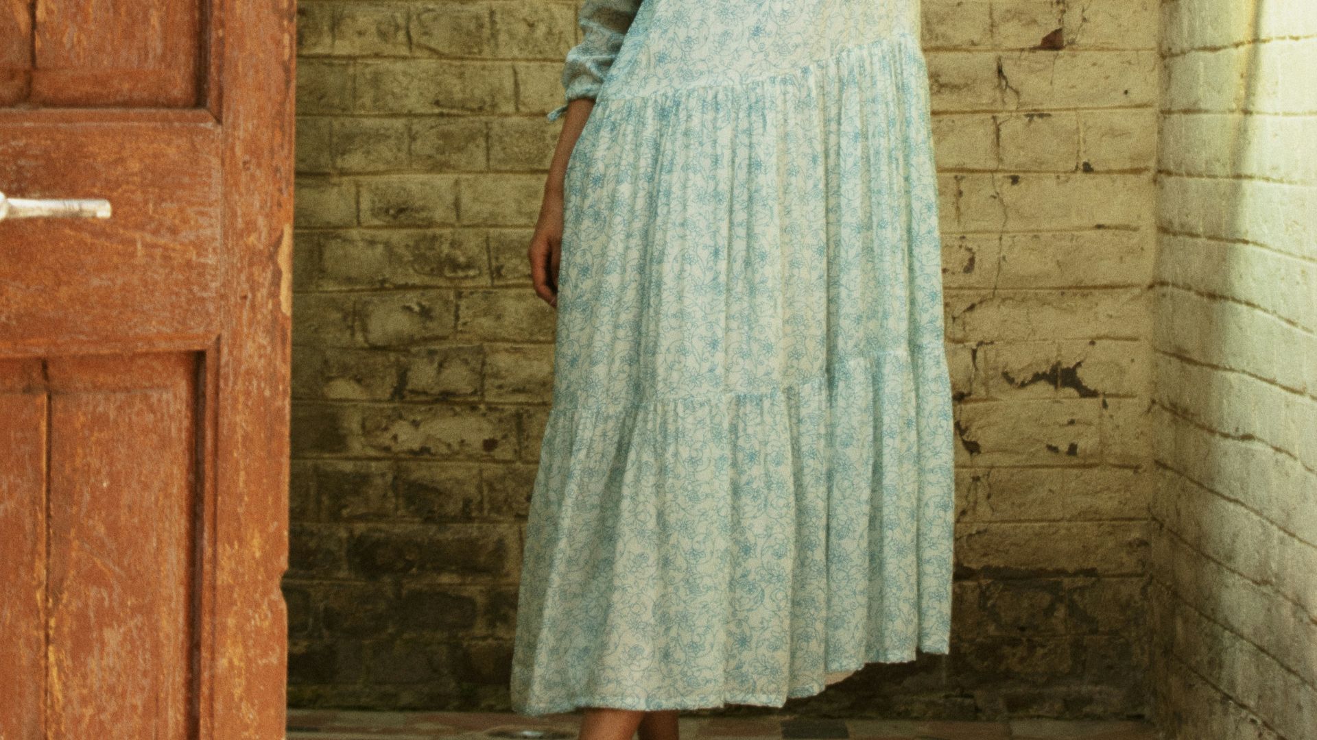 Young woman in a light blue dress leans against wall.