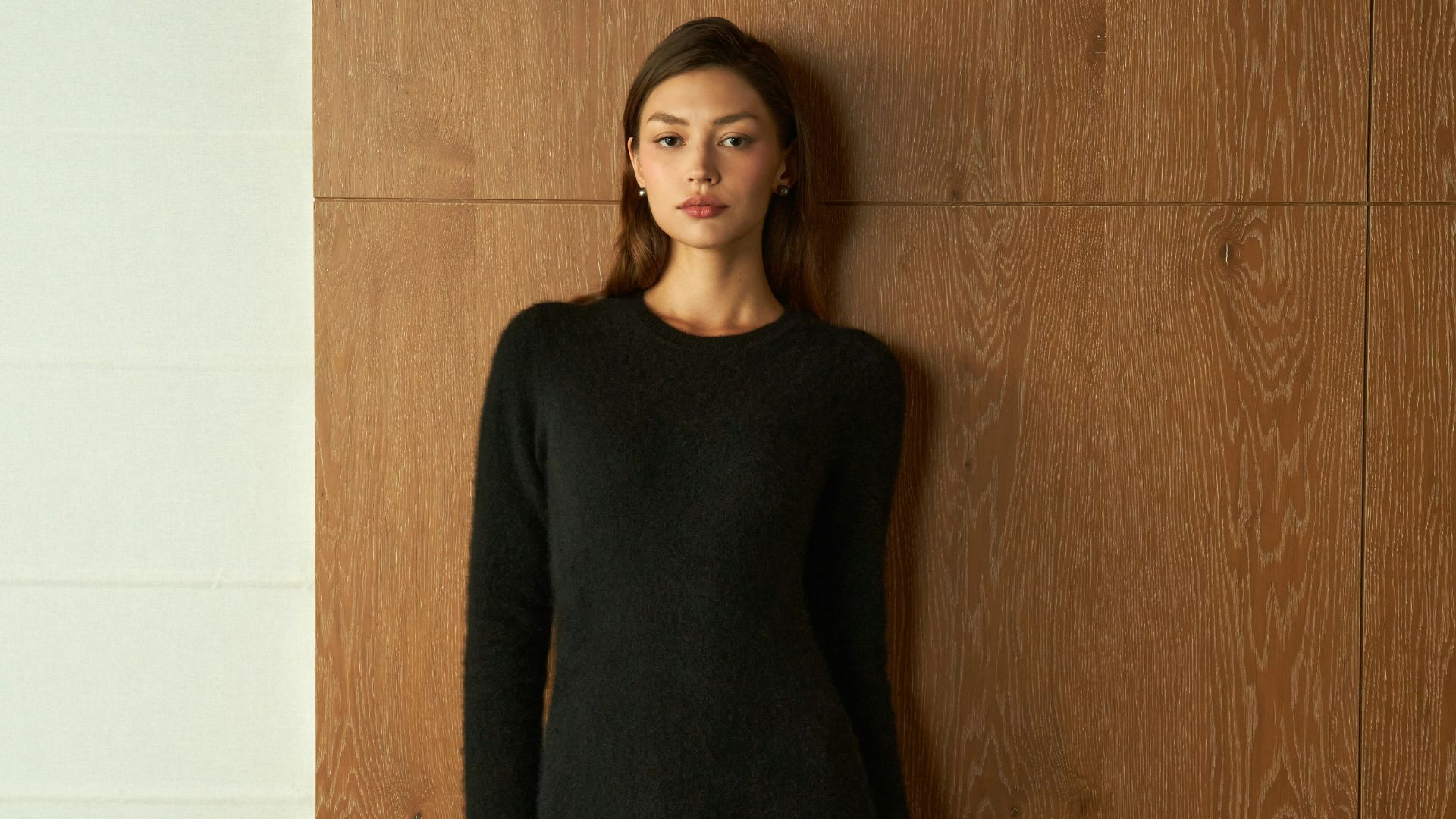 Young woman in a black dress standing against wood paneling.