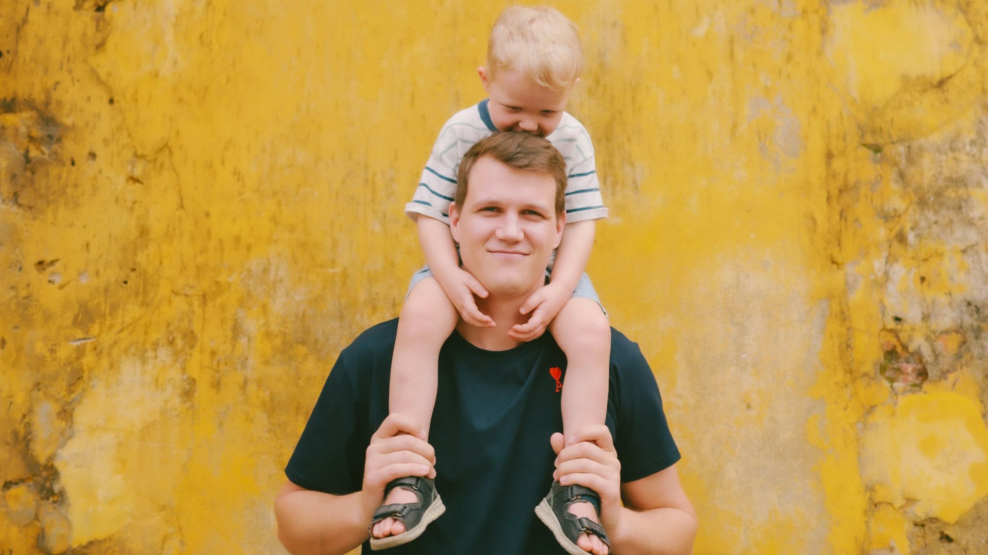 Father carries son on shoulders against yellow wall.