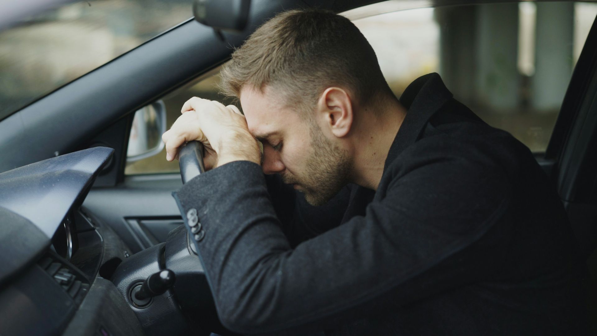 Man resting head on steering wheel in car.