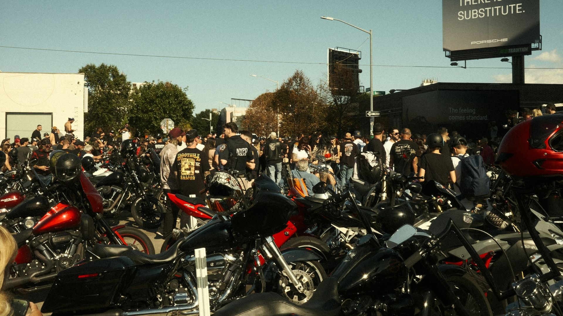 Crowd gathered around many motorcycles with a billboard.