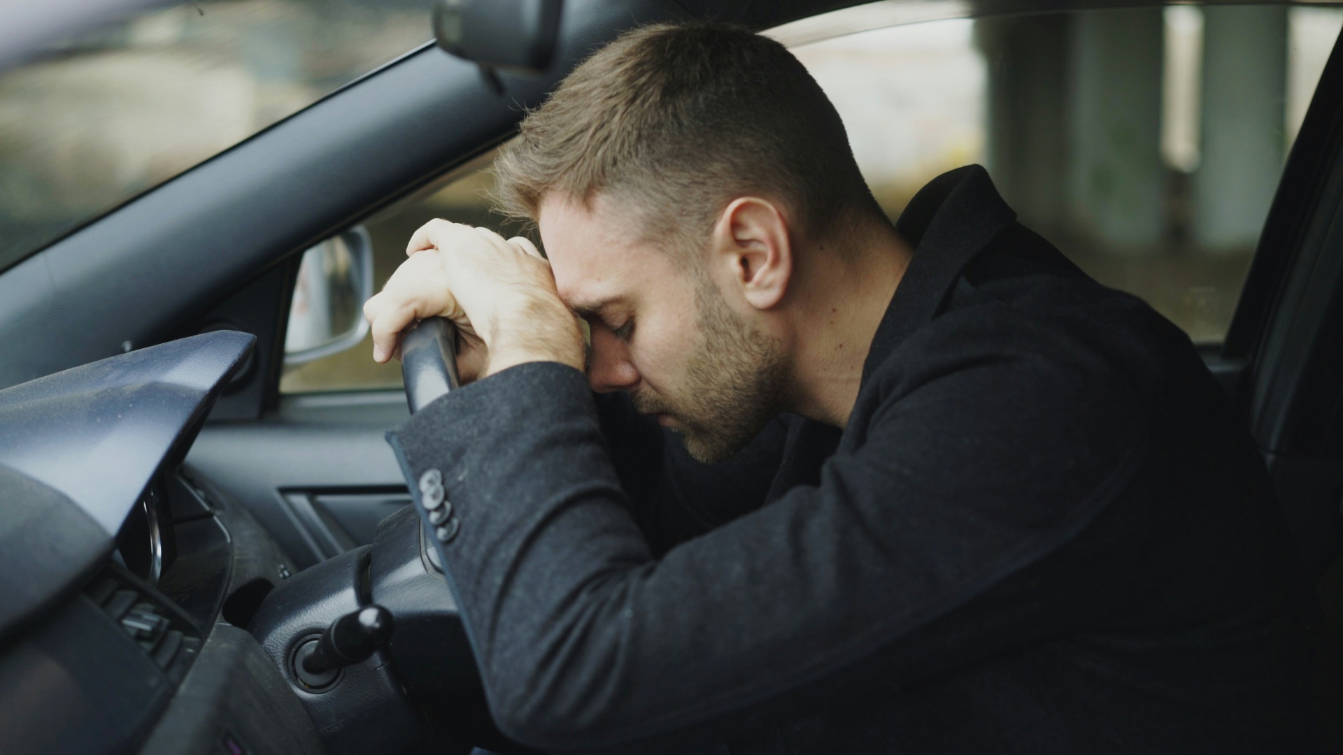 Man resting head on steering wheel in car.